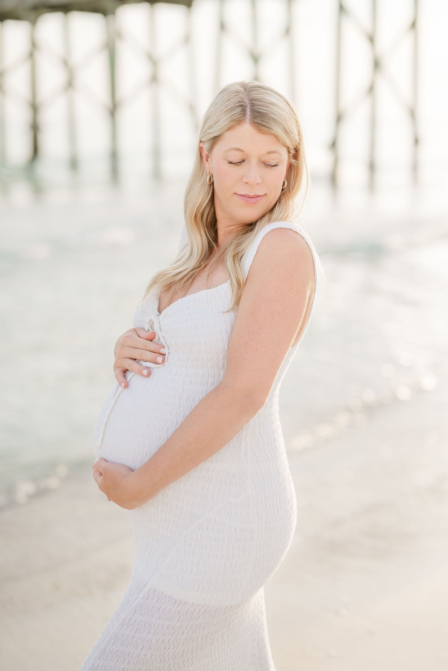 Expectant mother in white dress poses on sandy beach during golden hour in Panama City Beach, Fl..