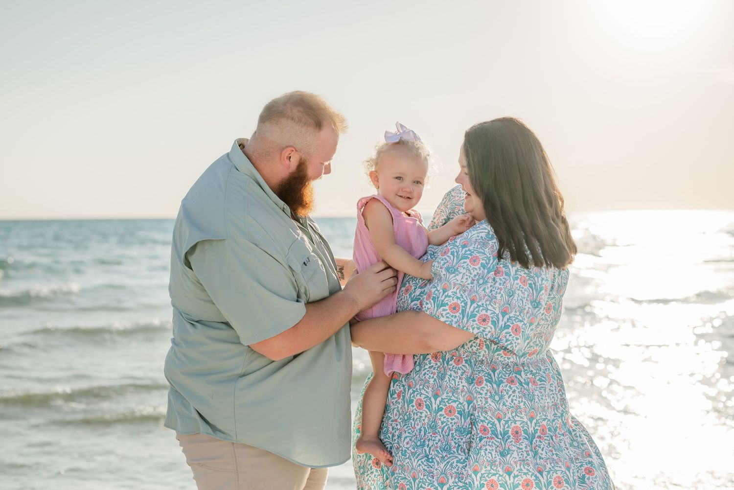 A couple holds their young child while standing on a beach at sunset.