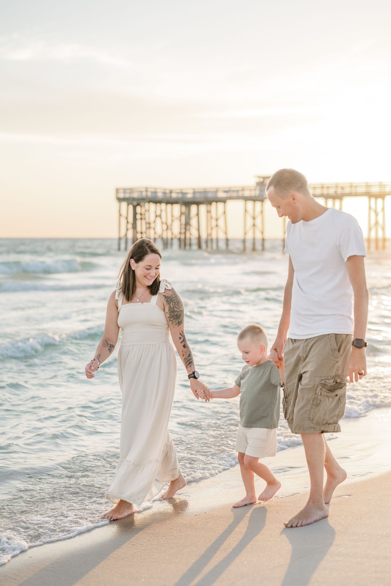 Family walking along beach at sunset with wooden pier in background.