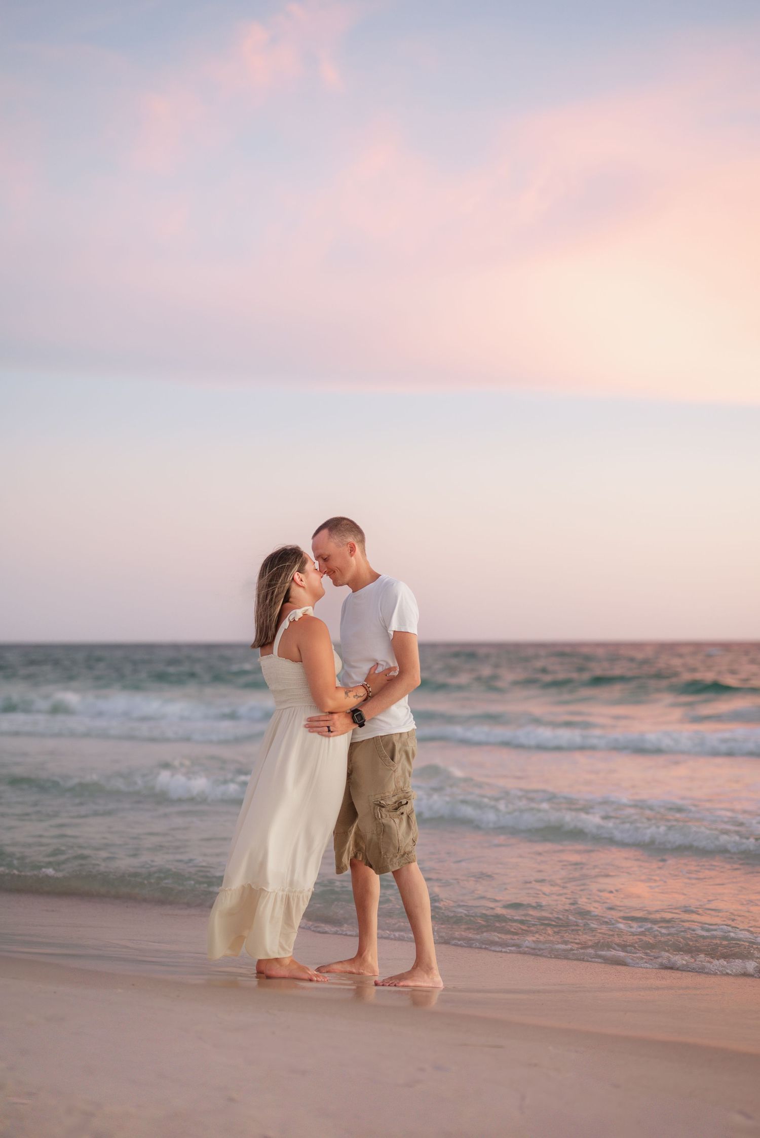 A couple embraces on a beach during sunset with soft pink skies and gentle waves in the background.