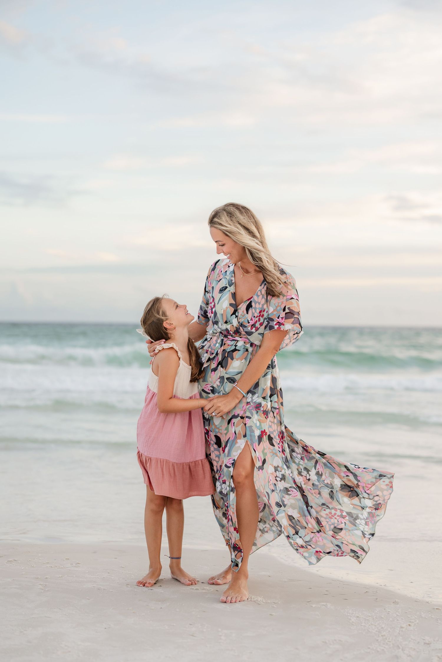 Mother and daughter in floral and pink dresses share playful moments on a beach at sunset.