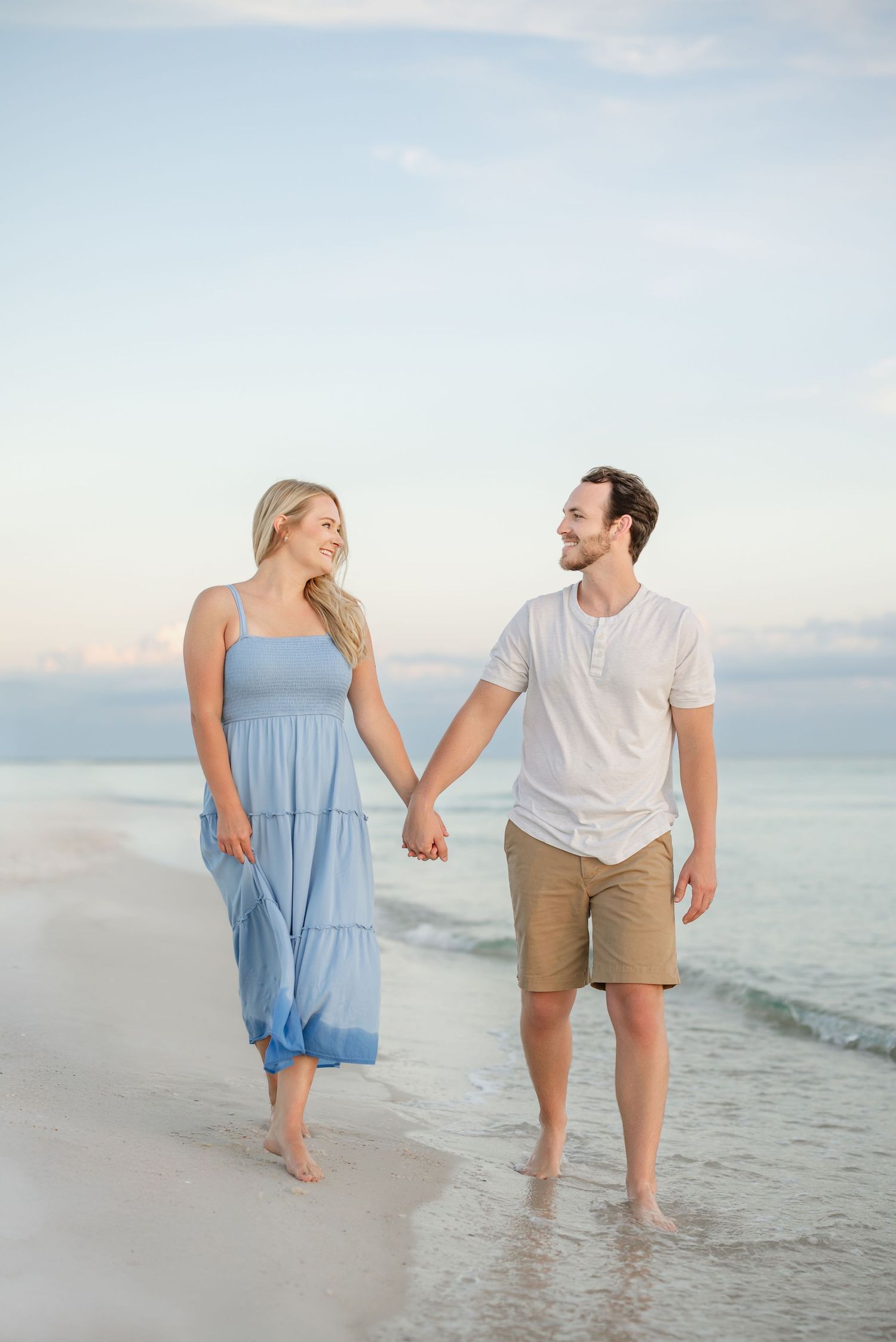 Couple in coordinated outfits walking along shoreline during sunset beach photoshoot.