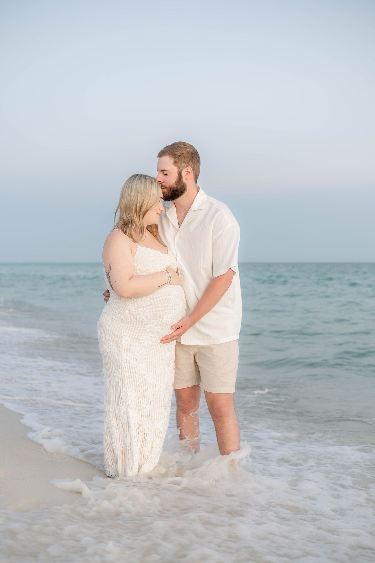 Romantic couple in white outfits embracing in ocean waves during sunset beach photoshoot.