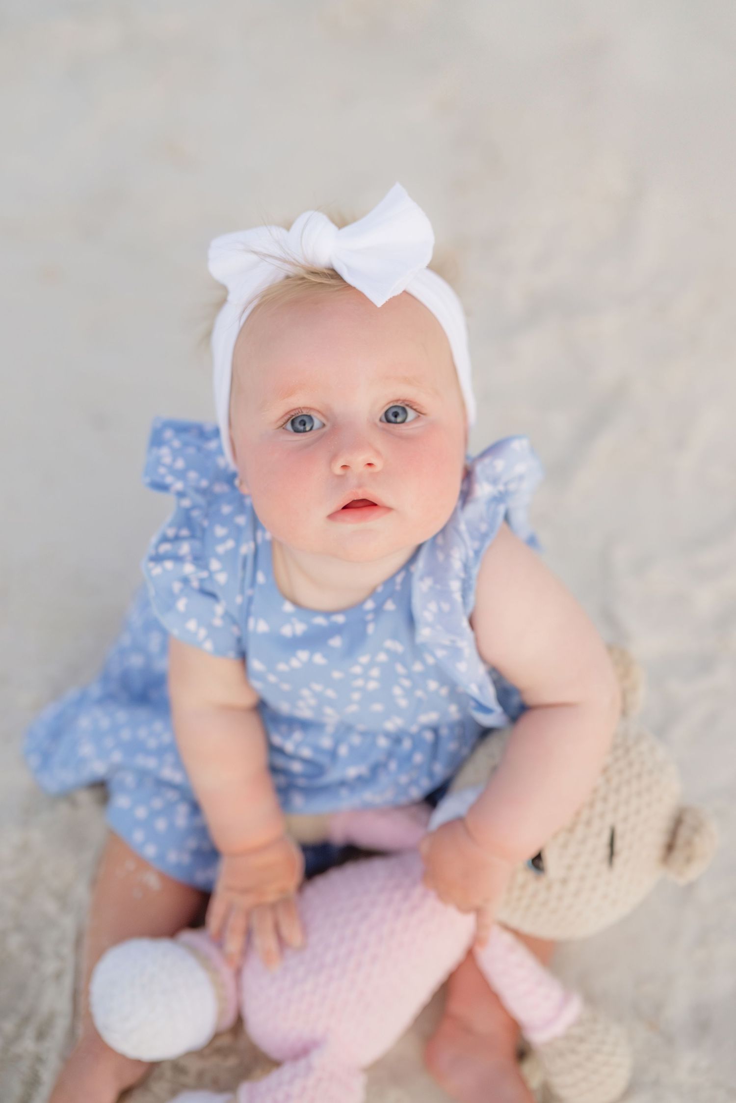 A baby wearing a blue floral romper and white headbow sits with a plush toy.