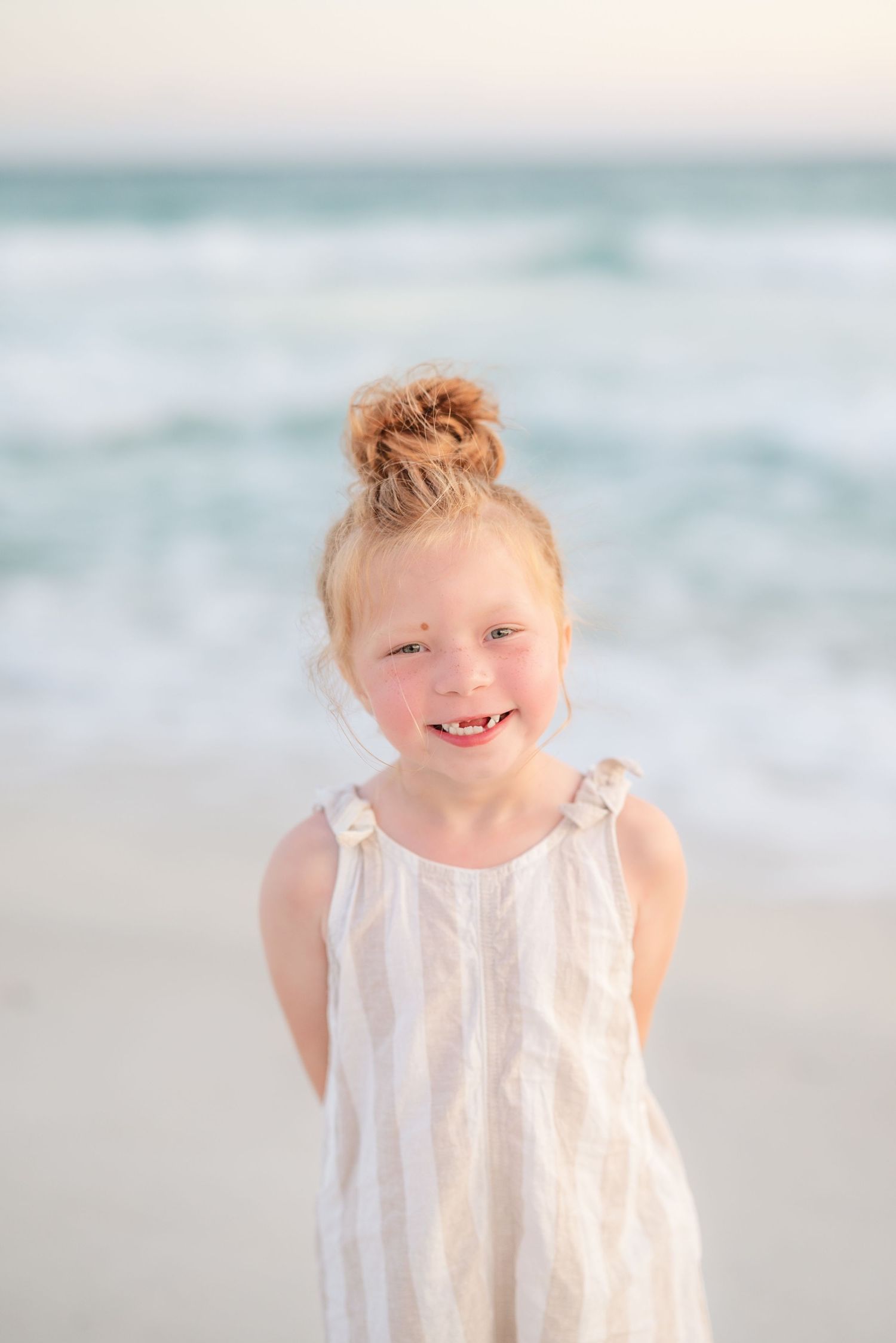 Young child in white dress smiling and playing on a sunny beach with ocean waves in background in Panama City Beach, Fl..