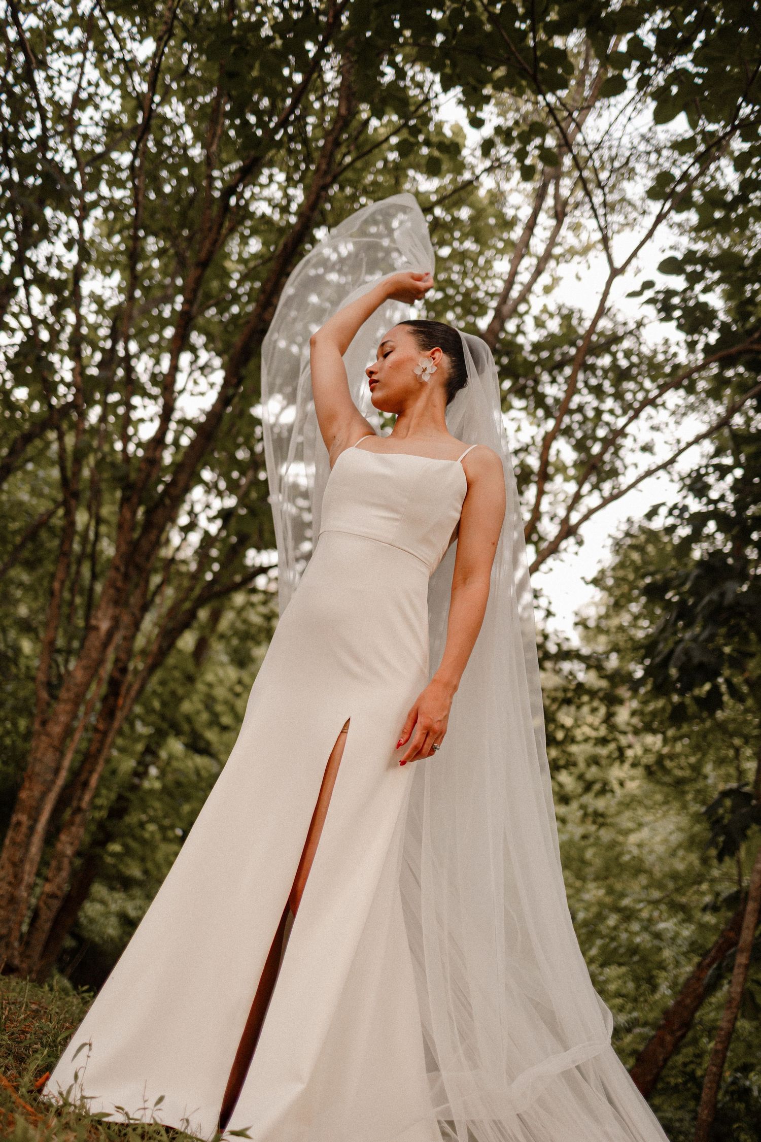 Elegant wedding dress with thigh slit and flowing veil in forest setting.