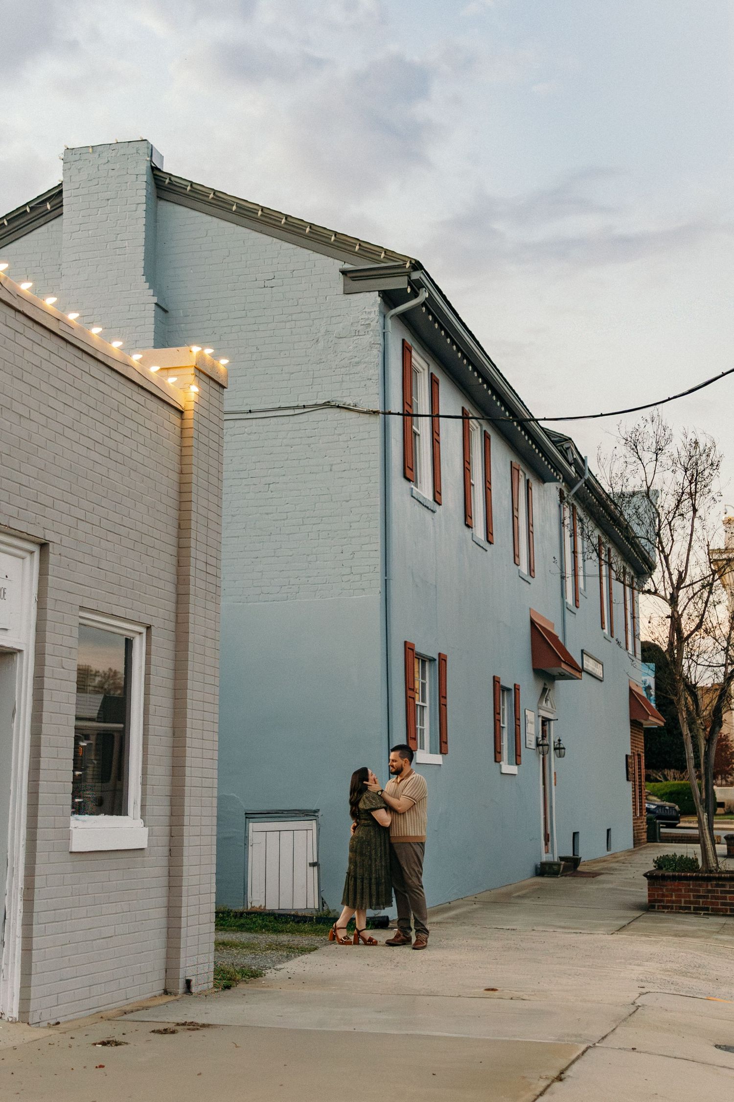 Couple embracing outside a blue brick building with string lights at dusk on a charming small town sidewalk.