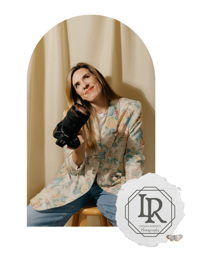 Professional photographer in blue floral top holds camera while posing against beige curtain backdrop.