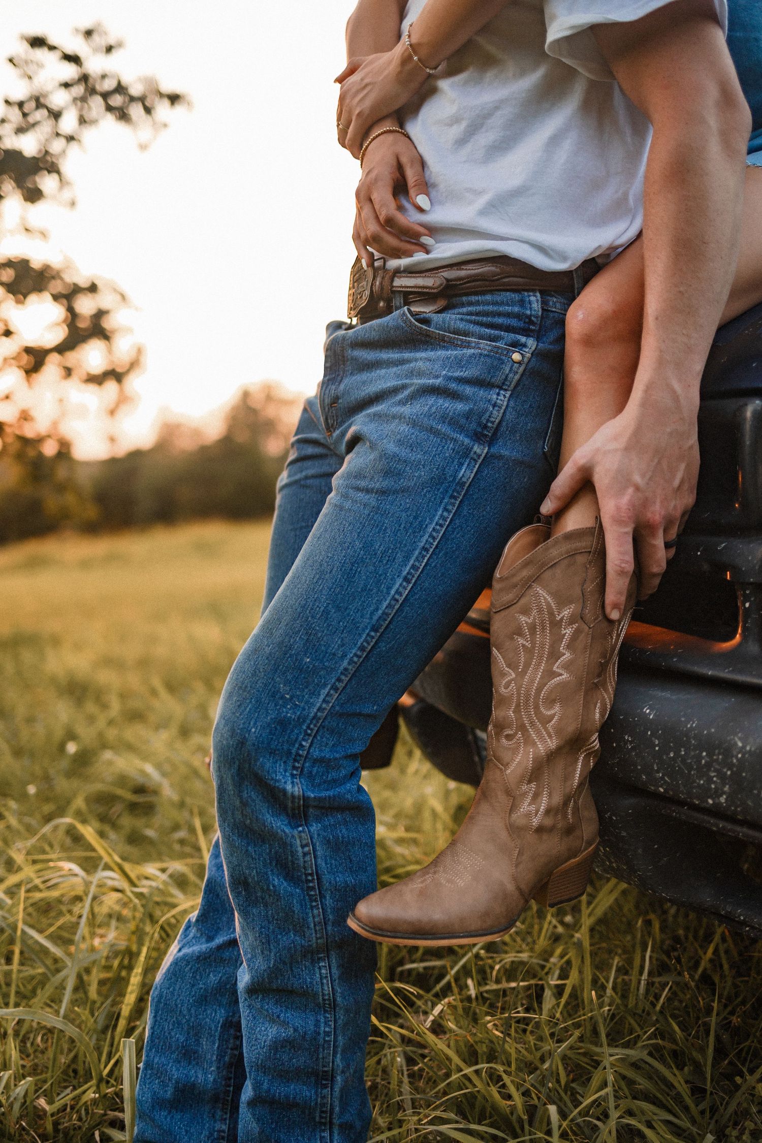Close-up view of brown cowboy boots and blue jeans against a truck tailgate at sunset in a grassy field.
