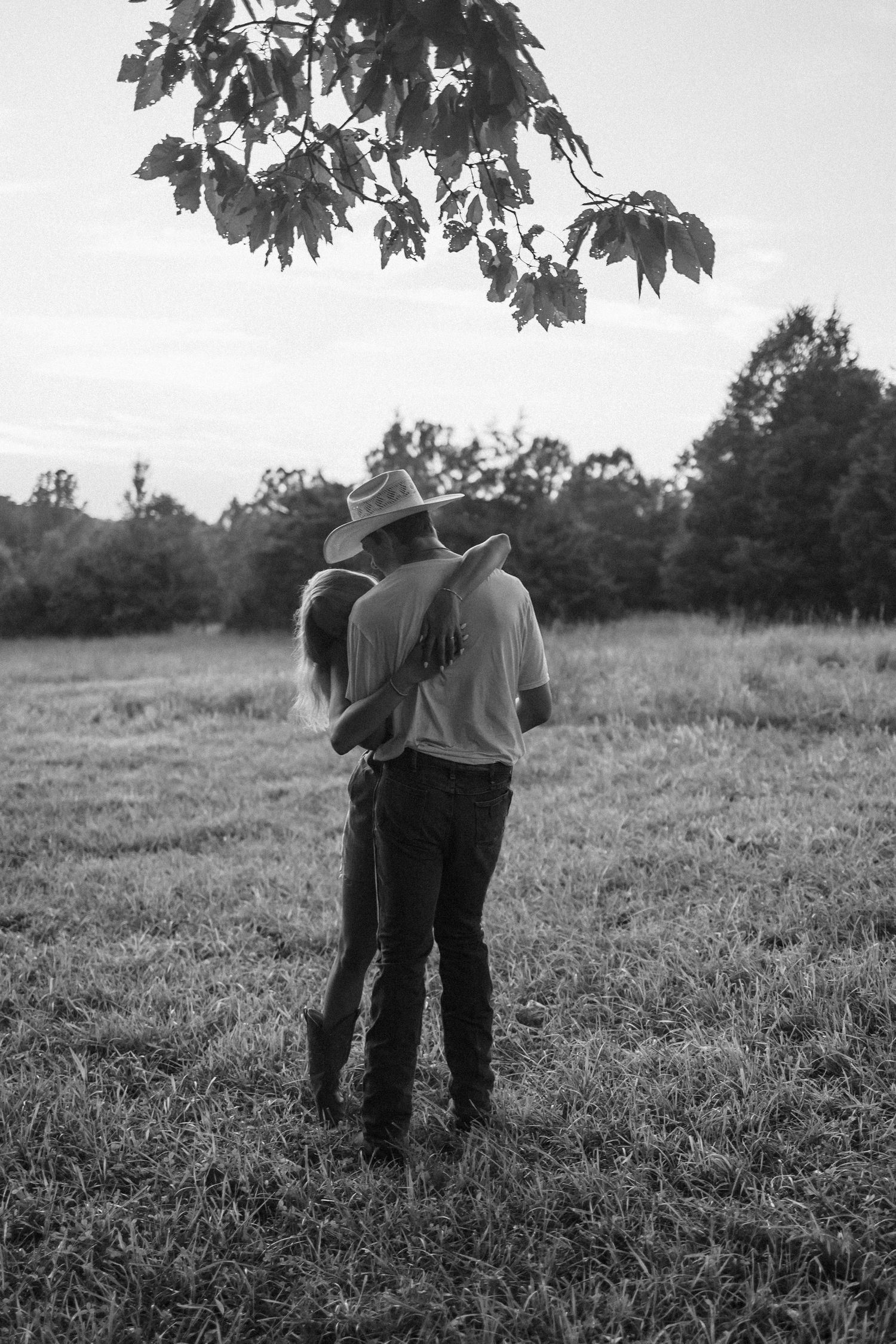 A black and white photo shows two people standing close together in a grassy field on a sunny day.