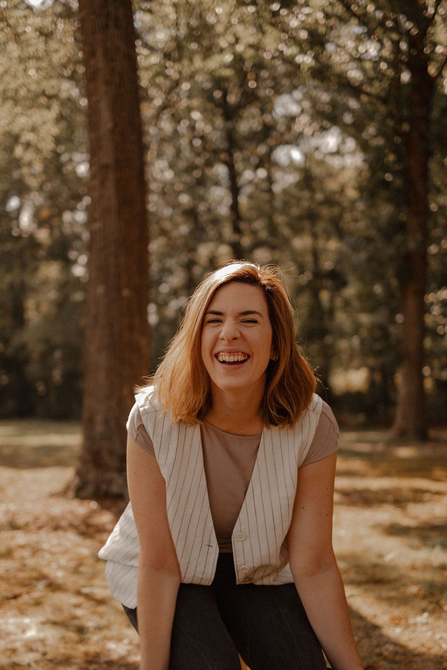 A woman in a white vest and brown top laughs joyfully while standing in a sunlit forest setting in Greensboro, NC.