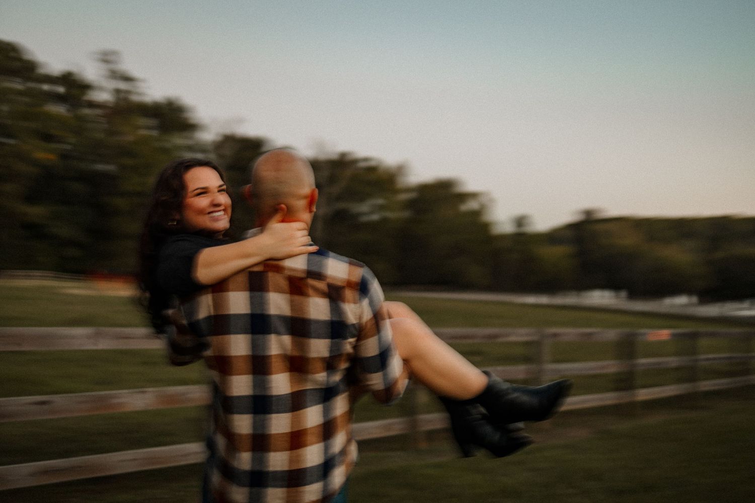 A man carrying his girlfriend in fast blurry motion with a field and fence in the background
