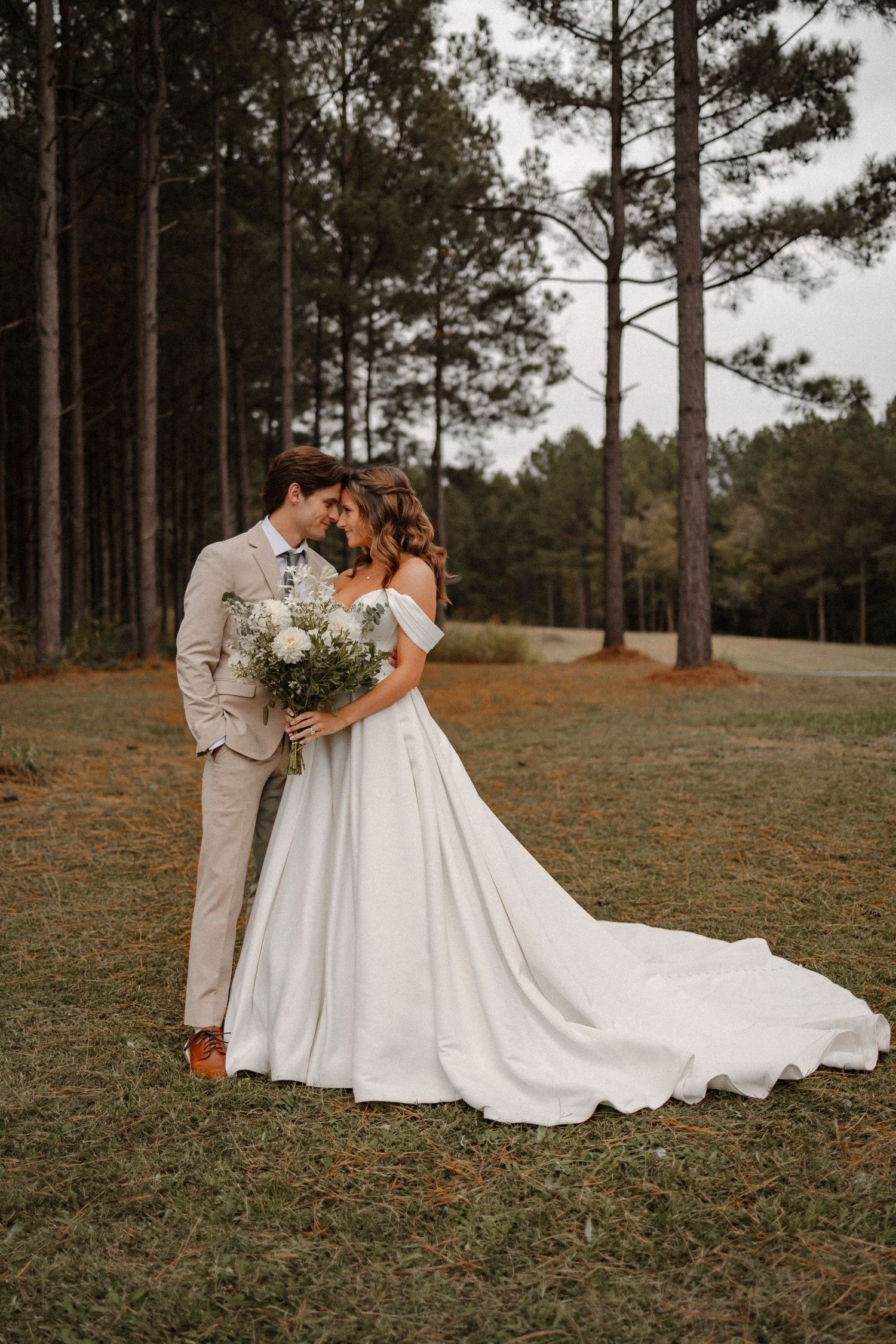 Bride in flowing white gown and groom in light suit embrace among tall pine trees.