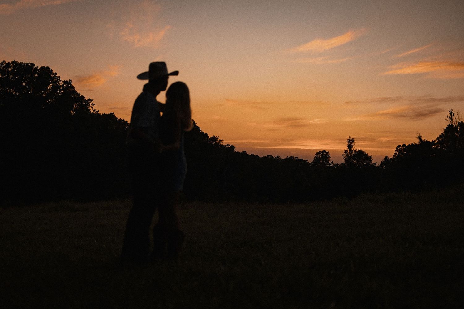 Silhouetted figures embrace while watching a dramatic orange and purple sunset over a rural landscape.