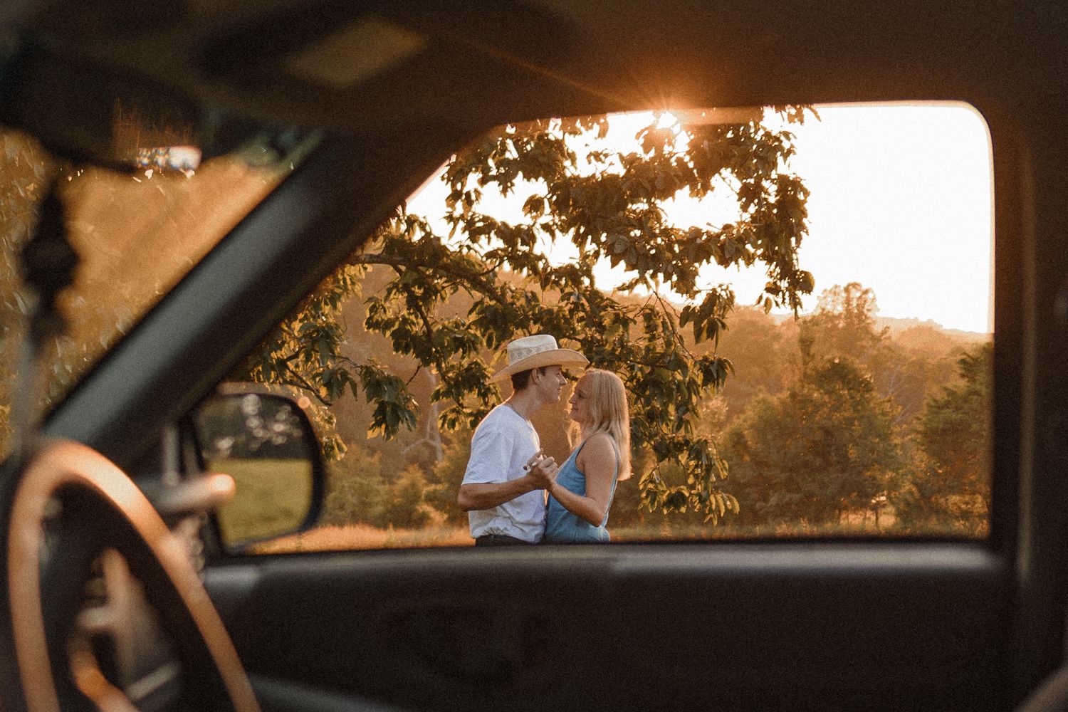 A romantic sunset scene viewed through a car window with two figures embracing in the warm golden light.