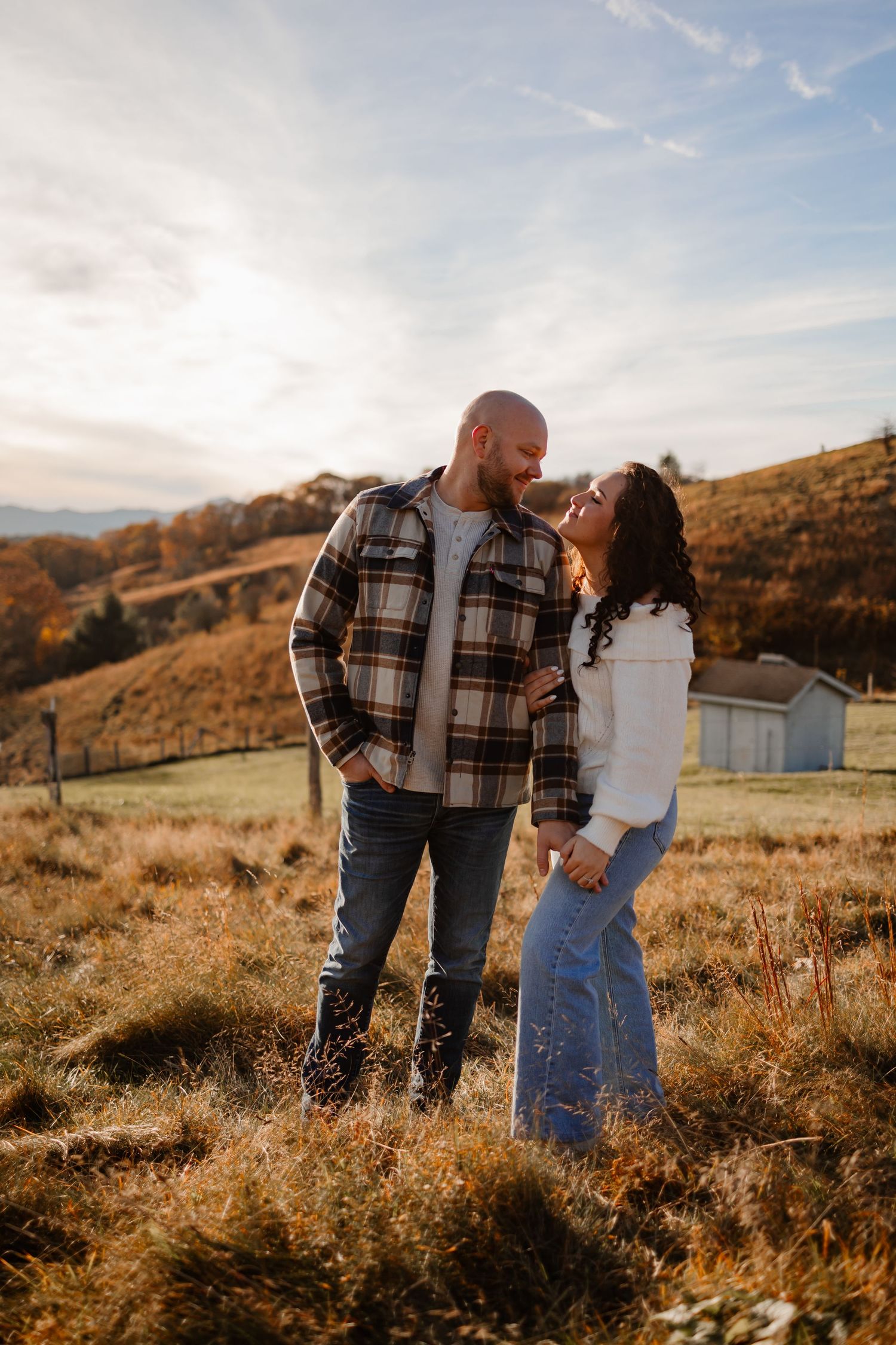 Couple in casual attire sharing tender moment during golden hour in rustic countryside setting.
