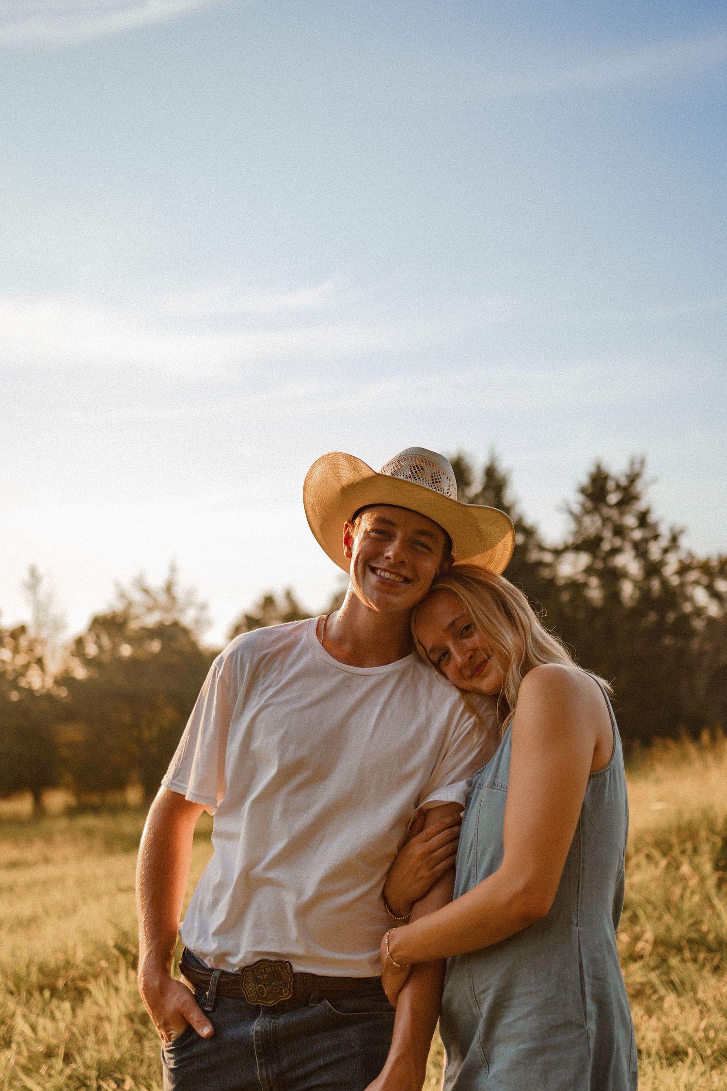 A couple embraces in a golden field at sunset while wearing casual clothes and a cowboy hat.