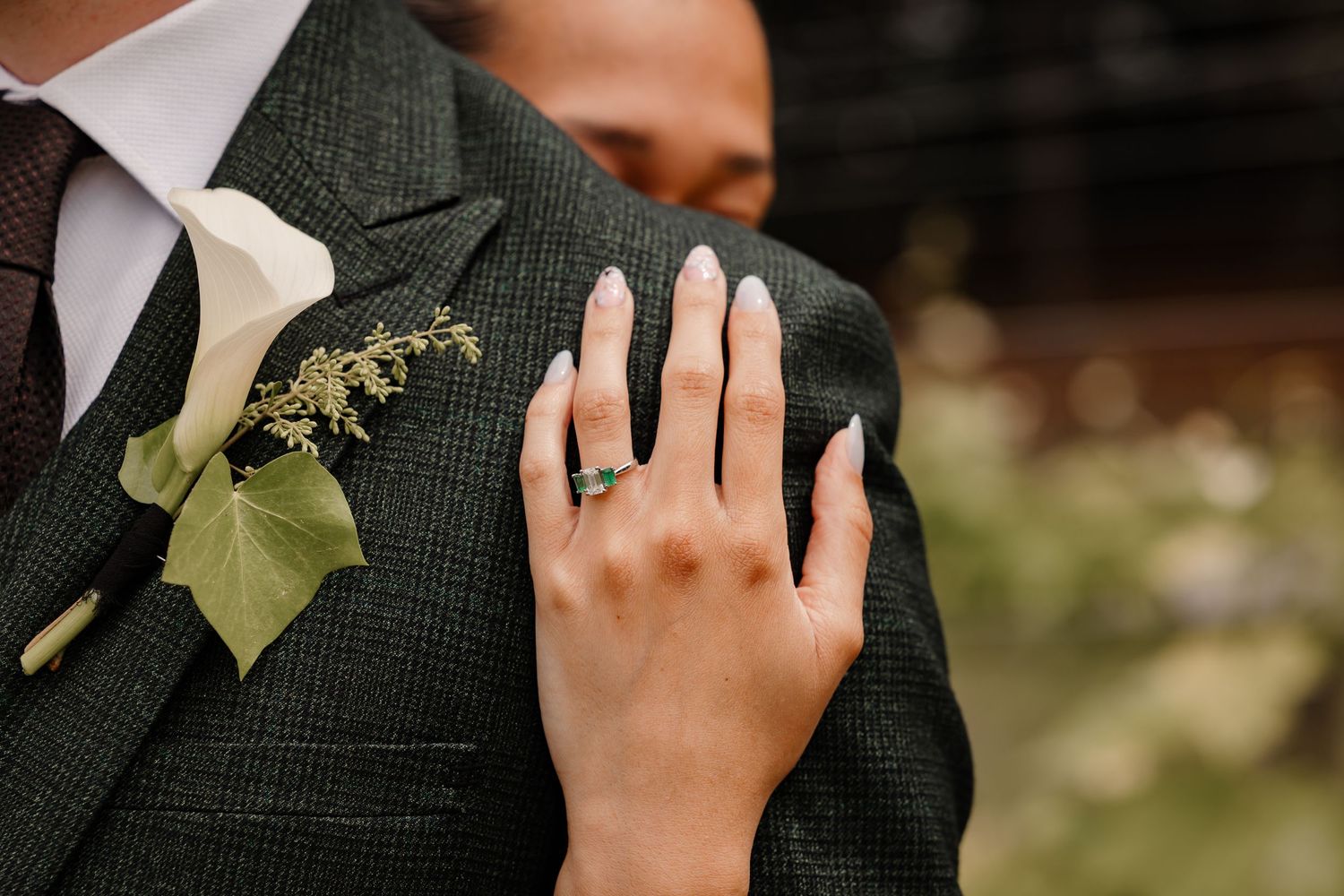 Close-up detail of a hand with ring resting on a dark green suit jacket with boutonniere.