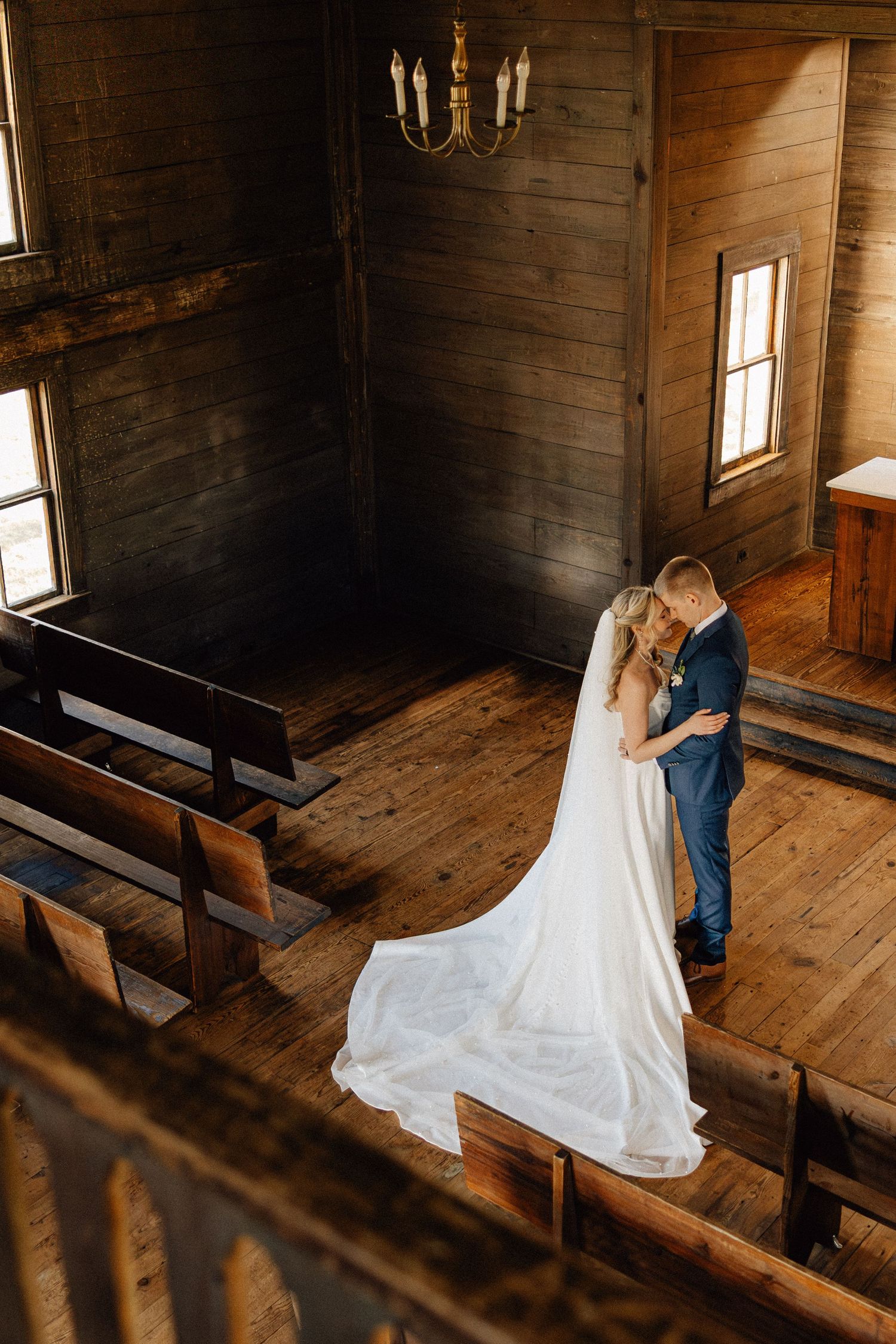 Bride and groom embracing in a rustic wooden chapel with pew seating, hardwood floors, and a chandelier overhead.