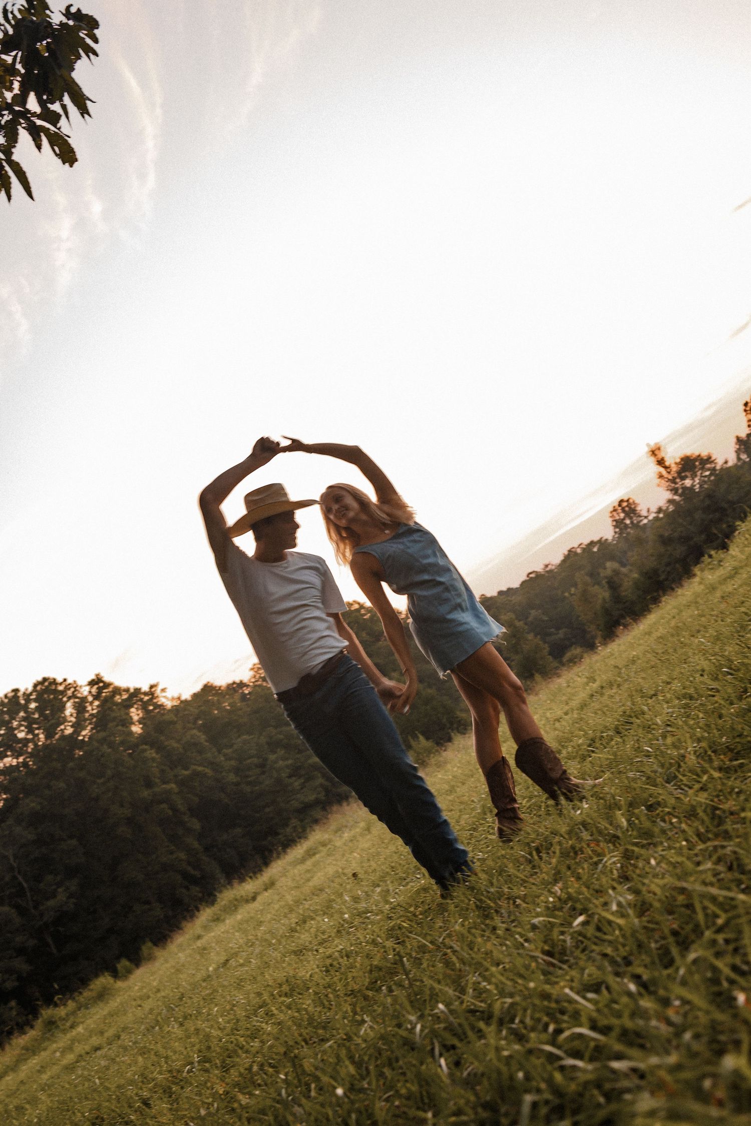 Two people holding hands while rolling down a grassy hillside at sunset in a playful moment.