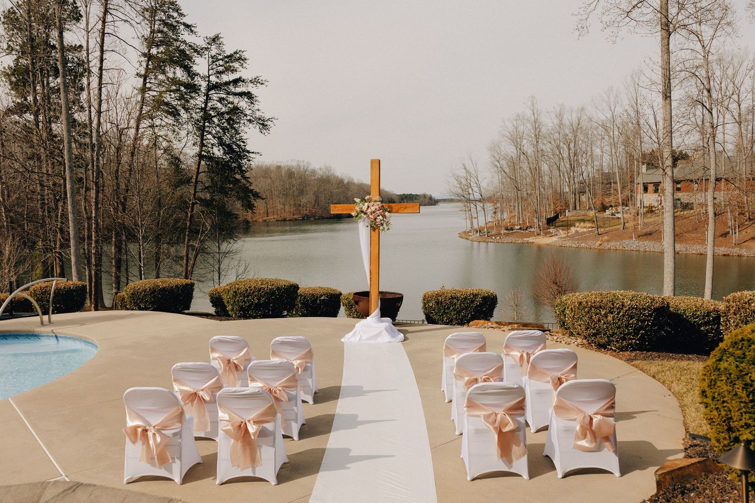 Lakeside wedding ceremony setup with white chairs and wooden cross at the altar.