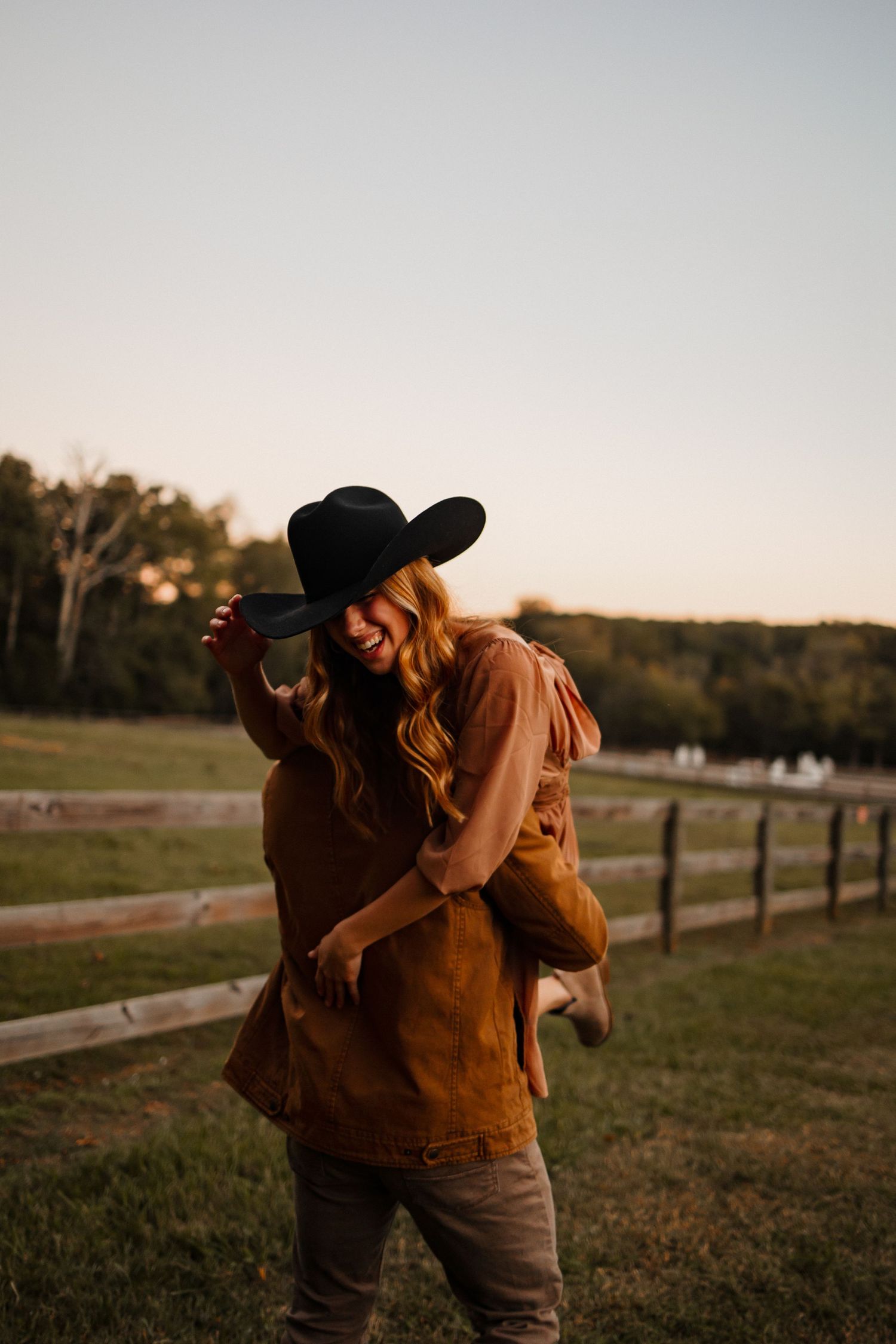 A girl with a cowboy hat on and getting carried over the back of her boyfriend in the country
