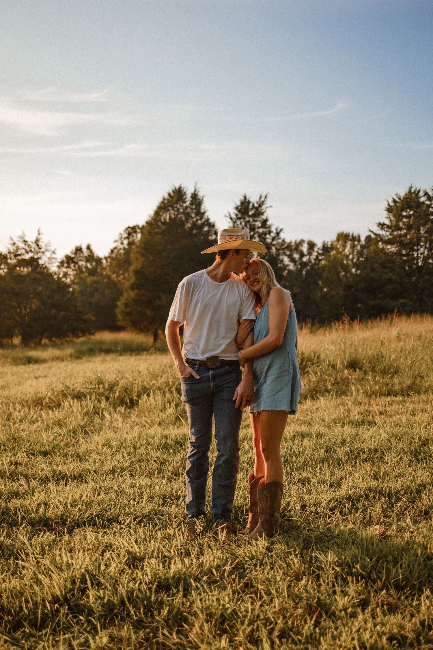 A couple stands together in a sunlit grassy meadow during golden hour as the sun sets behind pine trees.