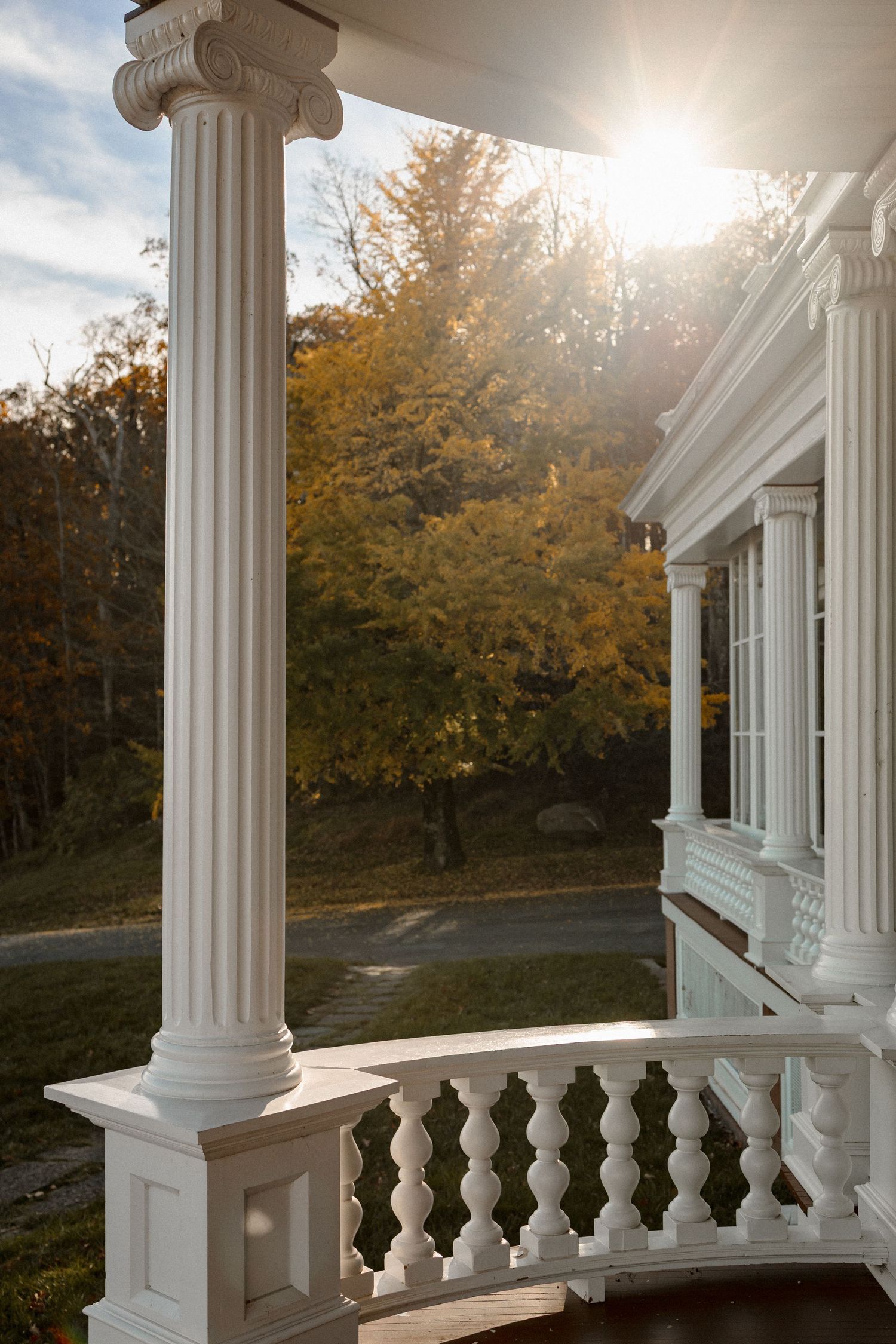 White classical columns and balustrade with autumn trees and sunlight in background.