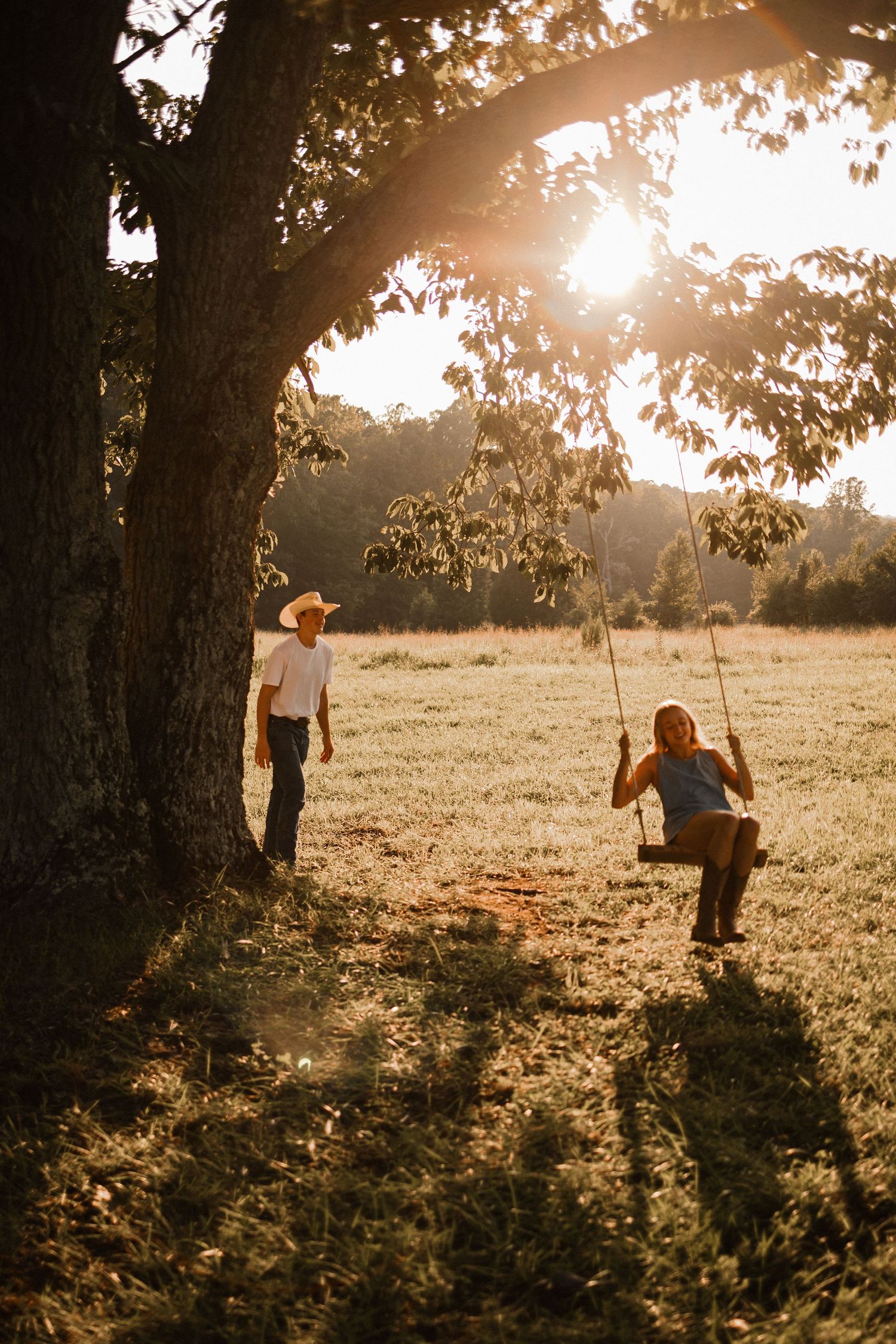 A person swings beneath a large tree at sunset creating a dreamy golden-hour silhouette in a peaceful meadow.