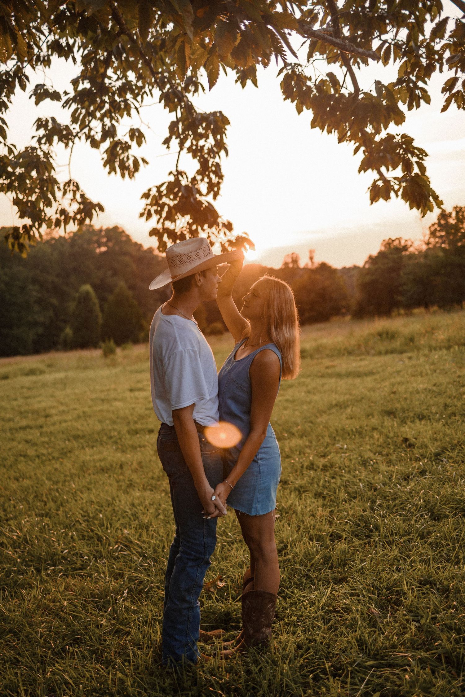 A couple shares an intimate moment in a grassy field at sunset with golden sunlight filtering through trees.