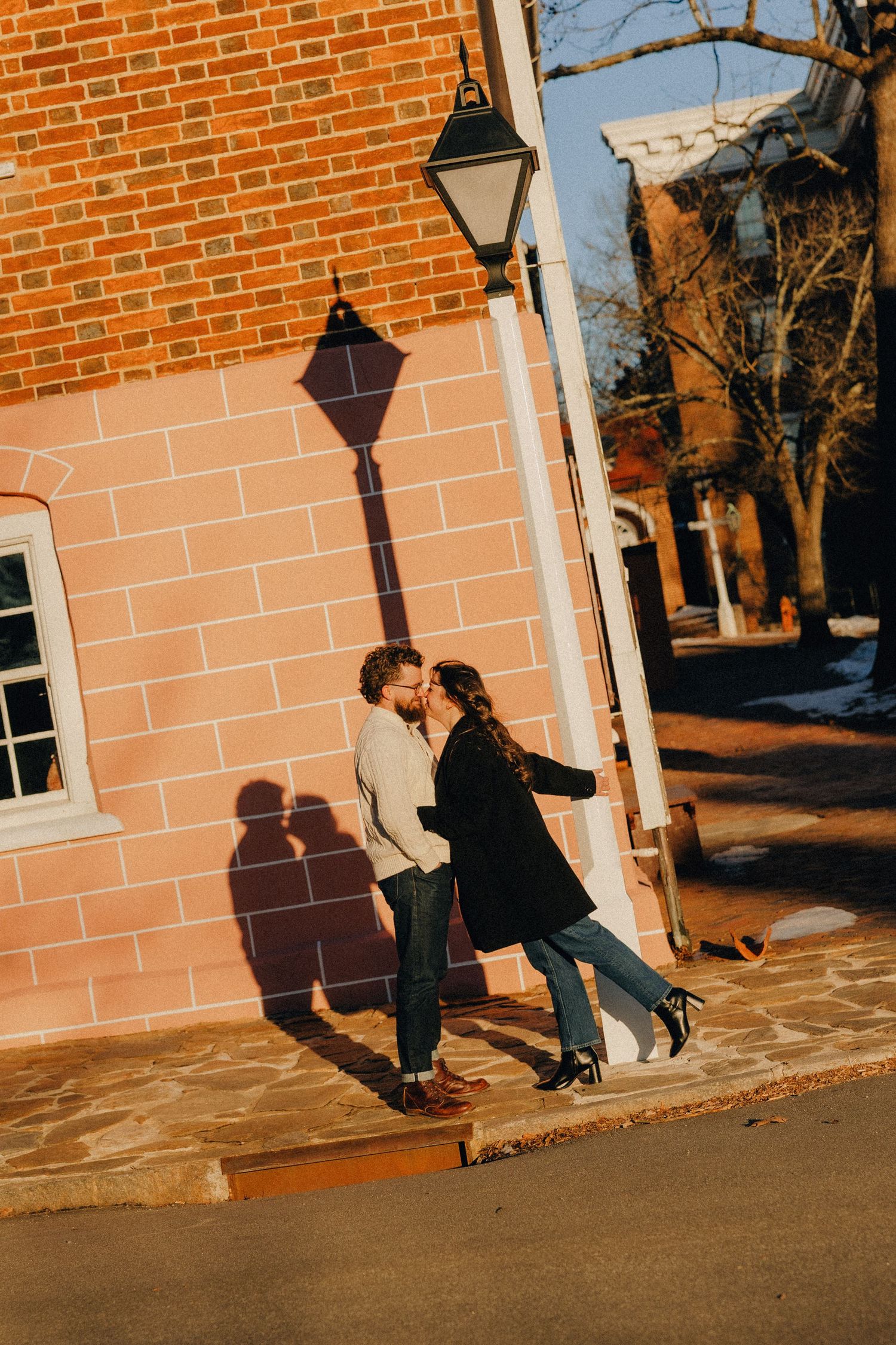 Couple embracing against brick wall with vintage street lamp casting romantic shadows at sunset.