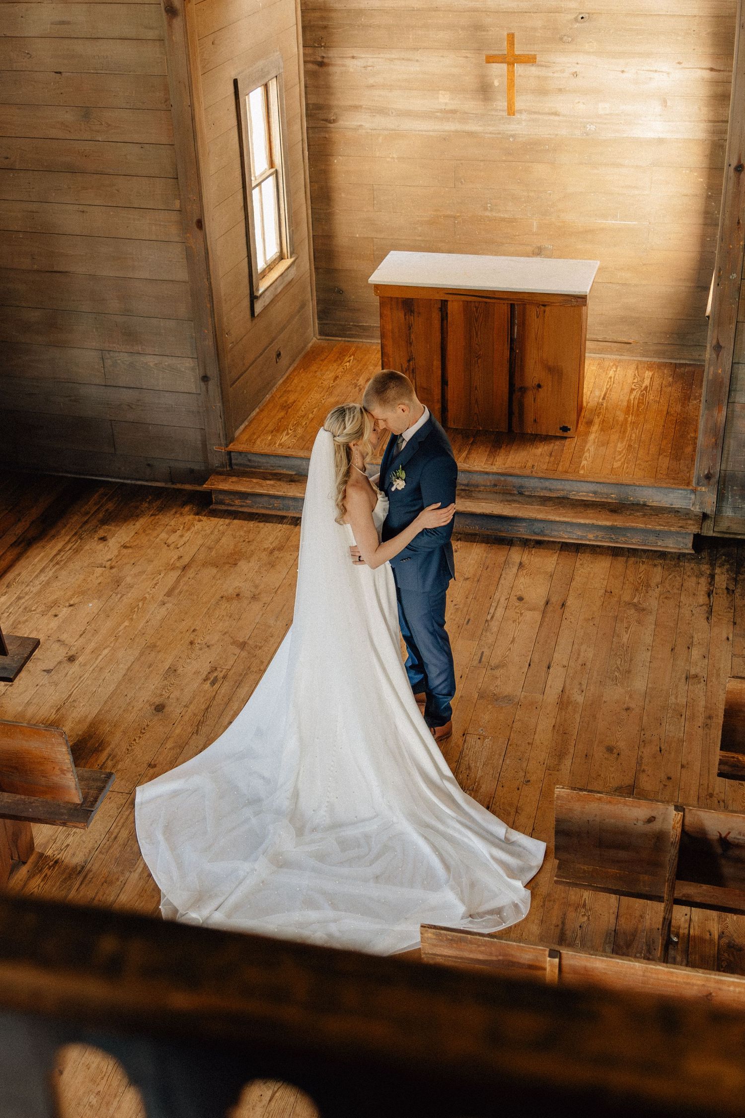 Bride and groom share a romantic embrace at the altar of a rustic wooden chapel with a cross on the wall.