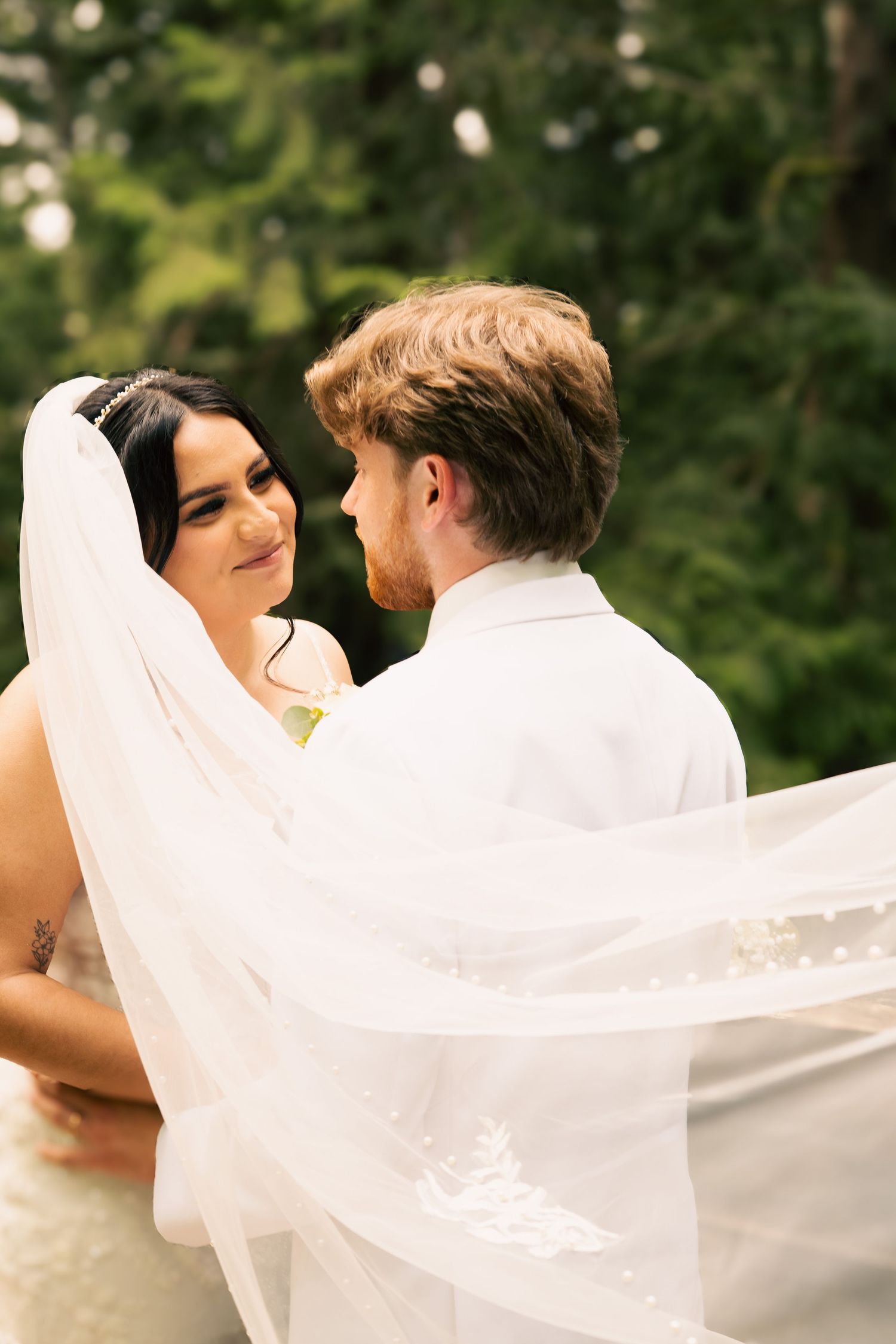 A couple stares lovingly into each others eyes. The bride wears a long sweeping veil.