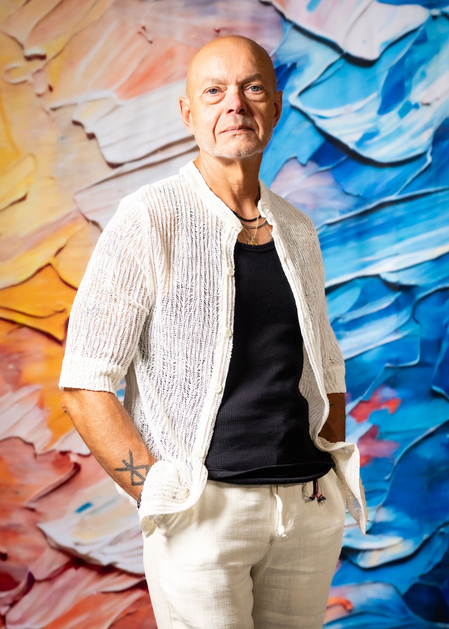 Bald man in white linen jacket and black shirt poses before colorful abstract painting backdrop.