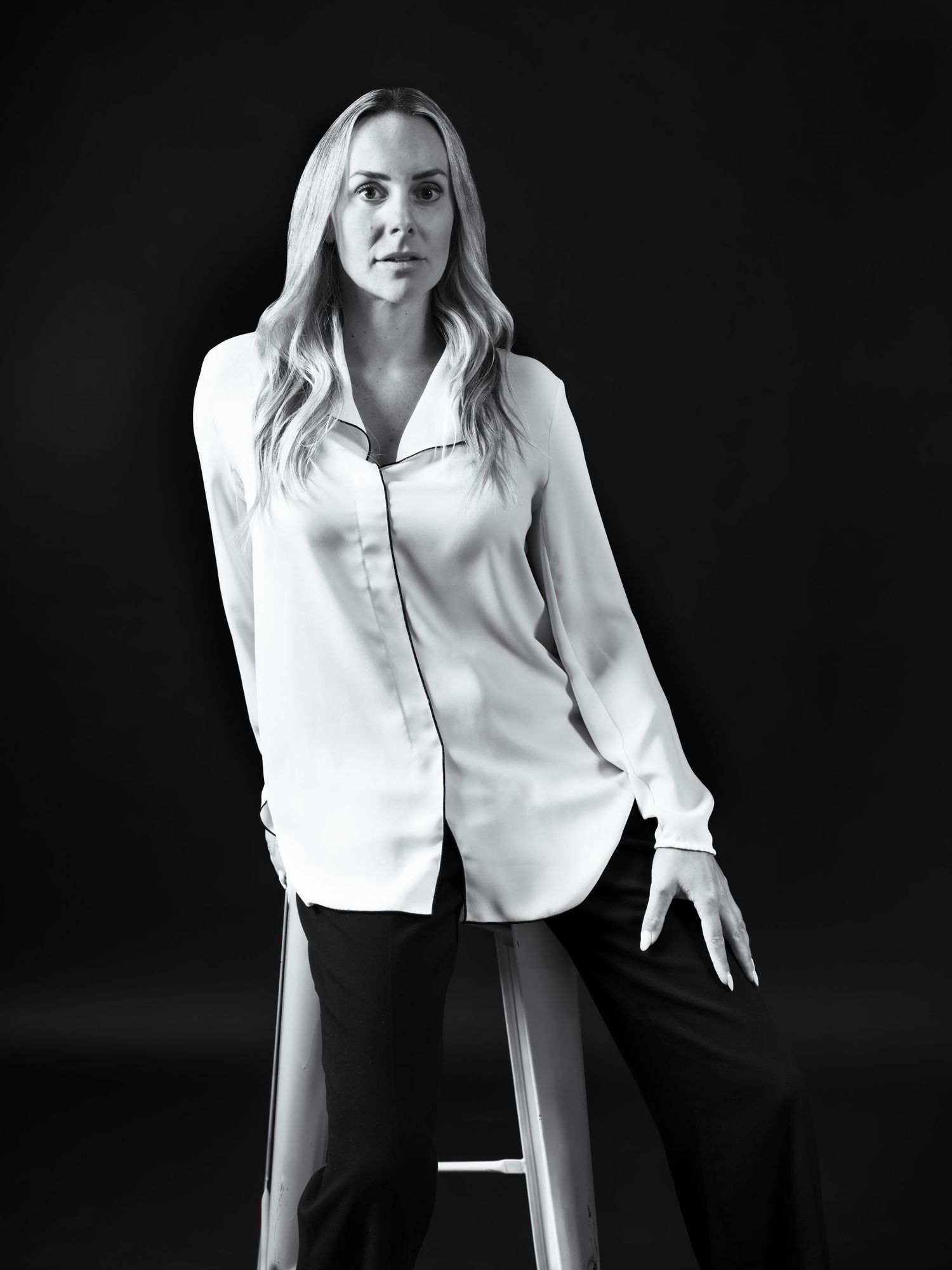 Black and white portrait of a blonde woman in a white silk blouse and black pants, seated on a stool.