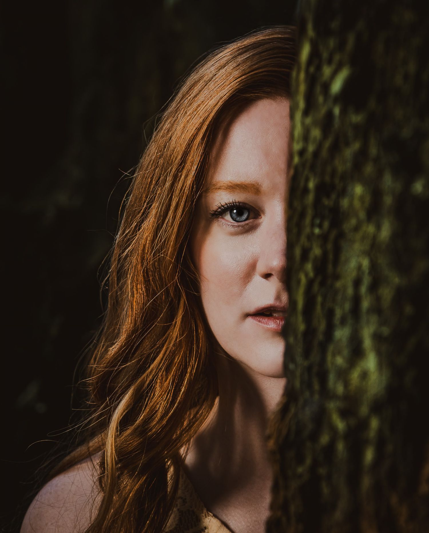 Close-up portrait of a red-haired woman with freckles peeking from behind a moss-covered tree.