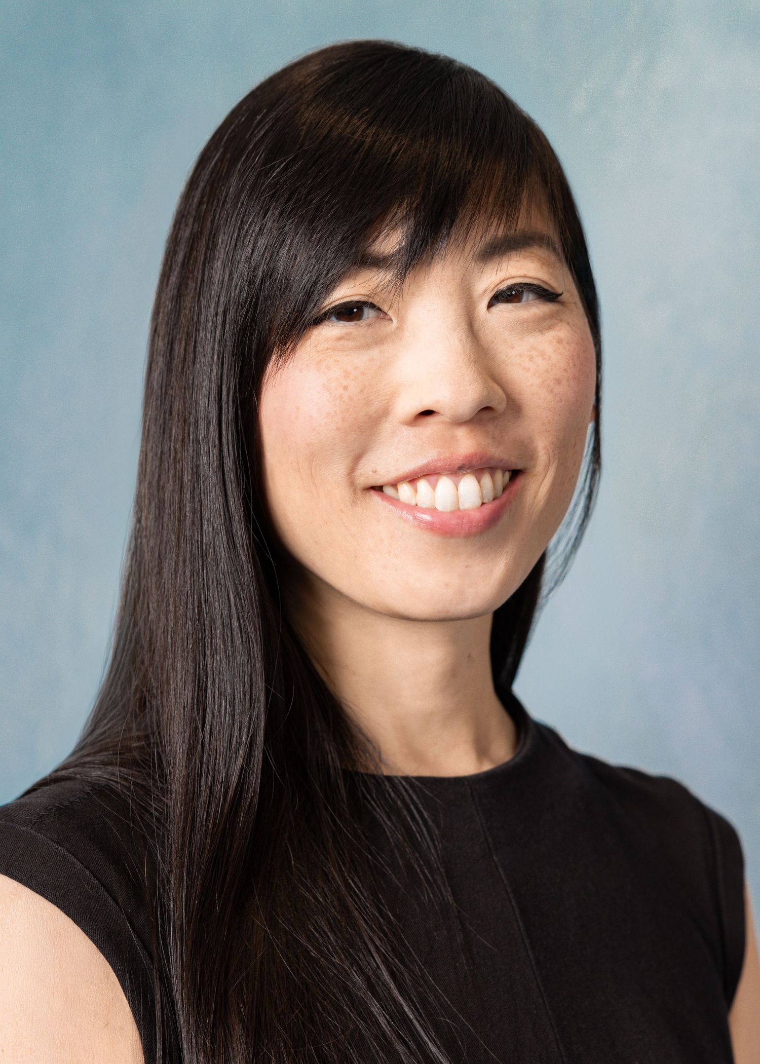 Smiling young Asian woman with long dark hair wearing a black top against a light blue background.