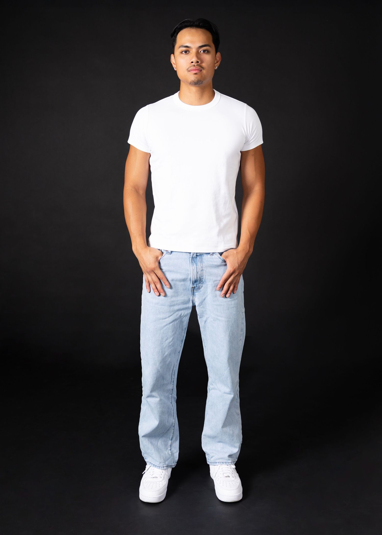 Man wearing a white fitted t-shirt and light wash baggy jeans with white sneakers posing against a dark background.