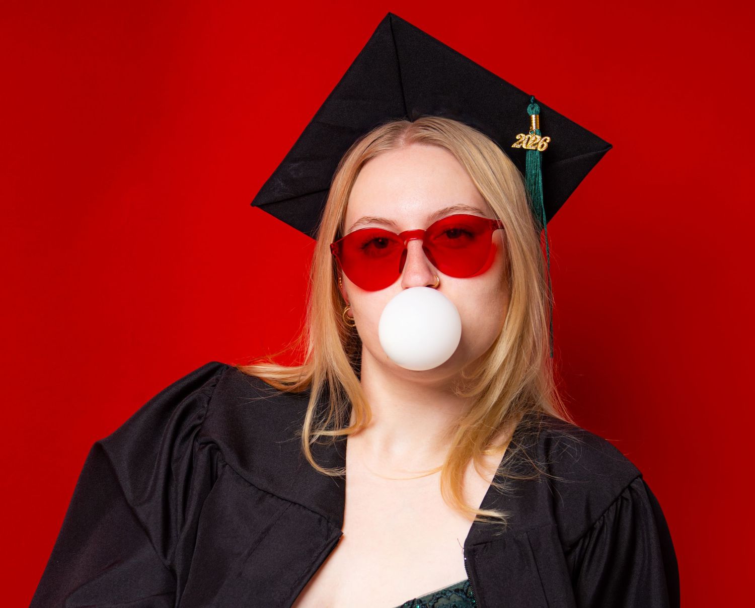 Young blonde woman in graduation cap, gown, and red sunglasses blowing a bubble gum bubble on red background.