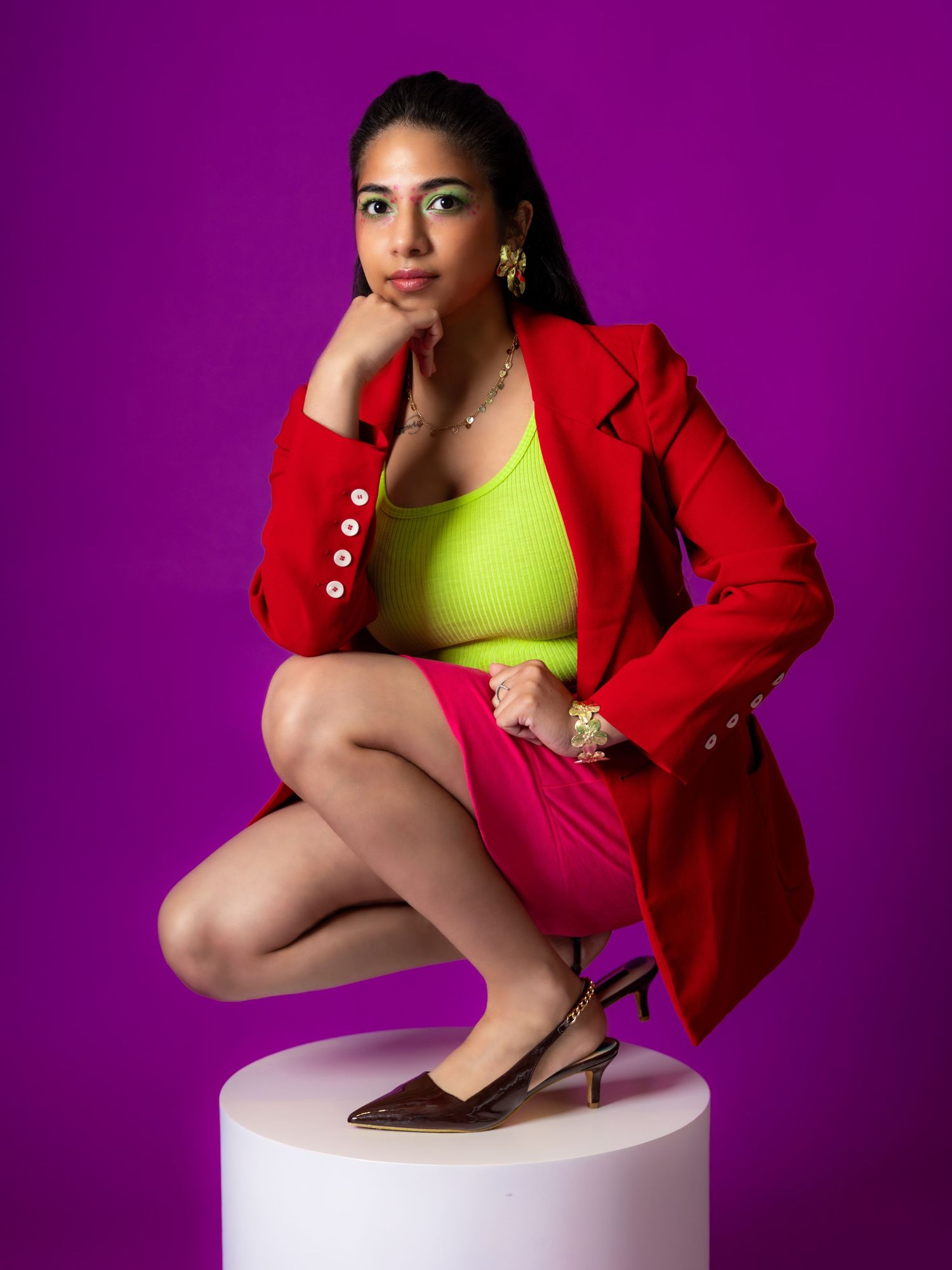 Woman in red blazer, neon top, and pink skirt posing on white pedestal against purple background.