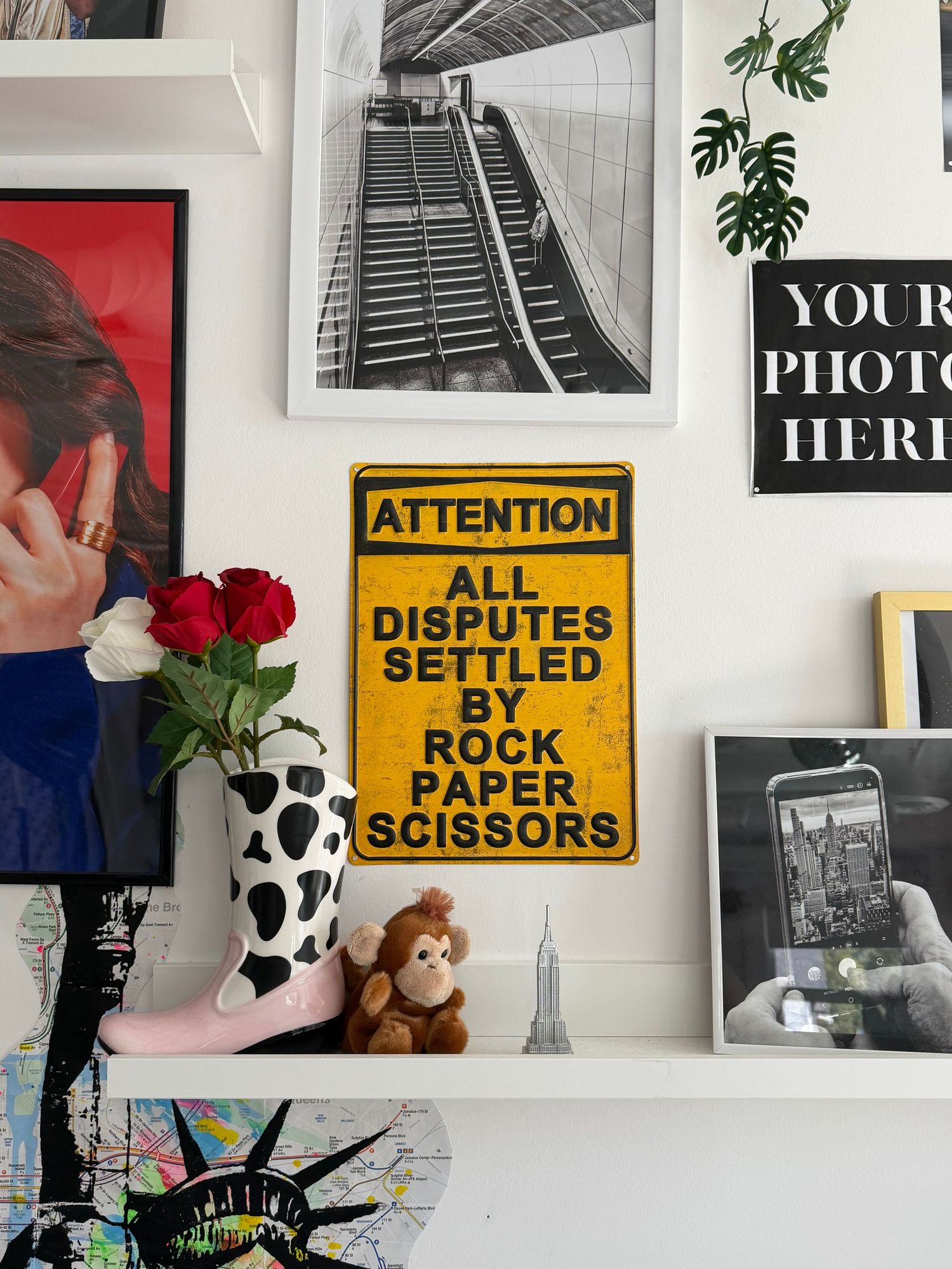 Yellow attention sign reading All Disputes Settled By Rock Paper Scissors displayed on a gallery wall with photos and decor.