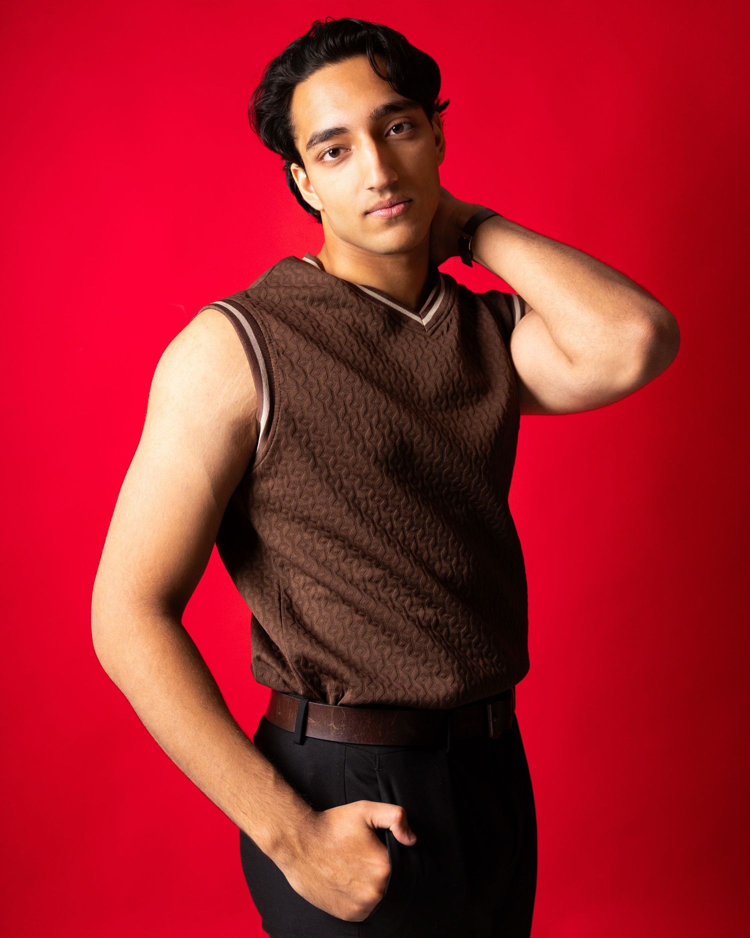 Young man in brown sleeveless sweater vest and black pants poses against vibrant red background.