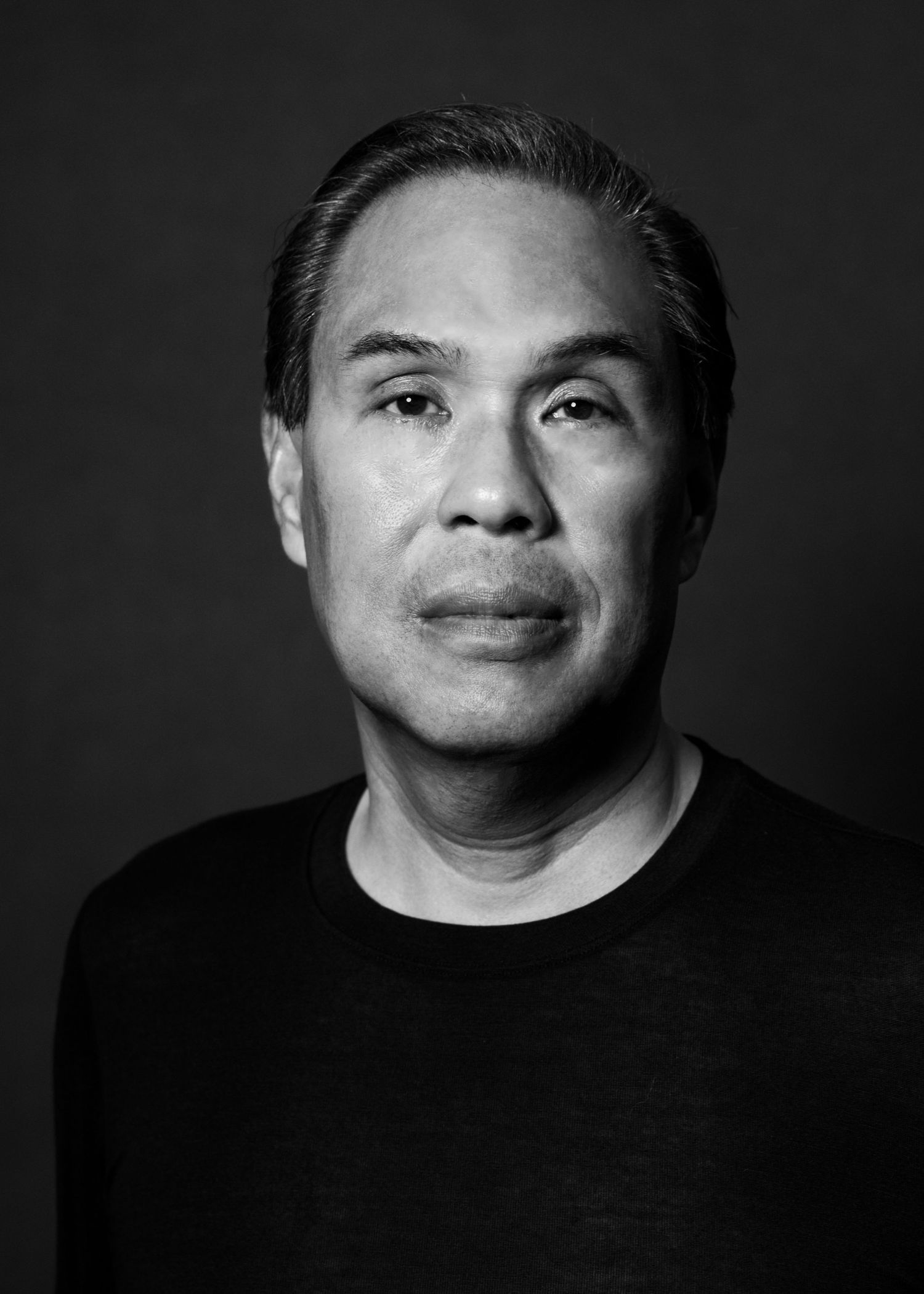 Black and white portrait of a middle-aged Asian man in a dark sweater against a dark background.