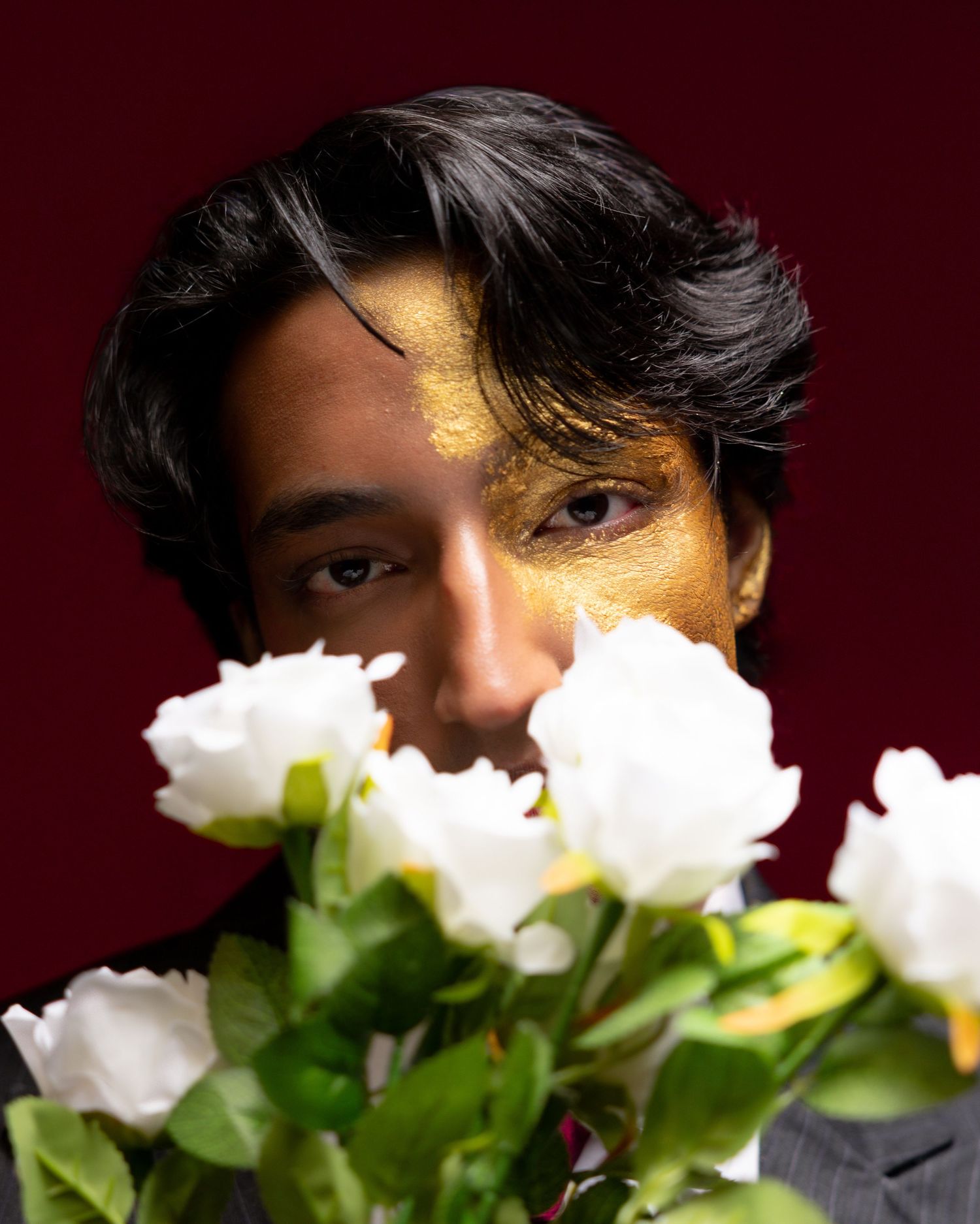 Person with gold face paint holding white roses against a dark red background.