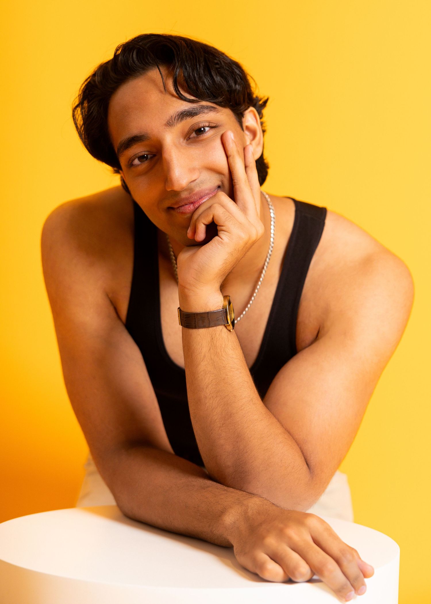 Young man in black tank top poses confidently against yellow background, wearing silver chain necklace and brown bracelet.