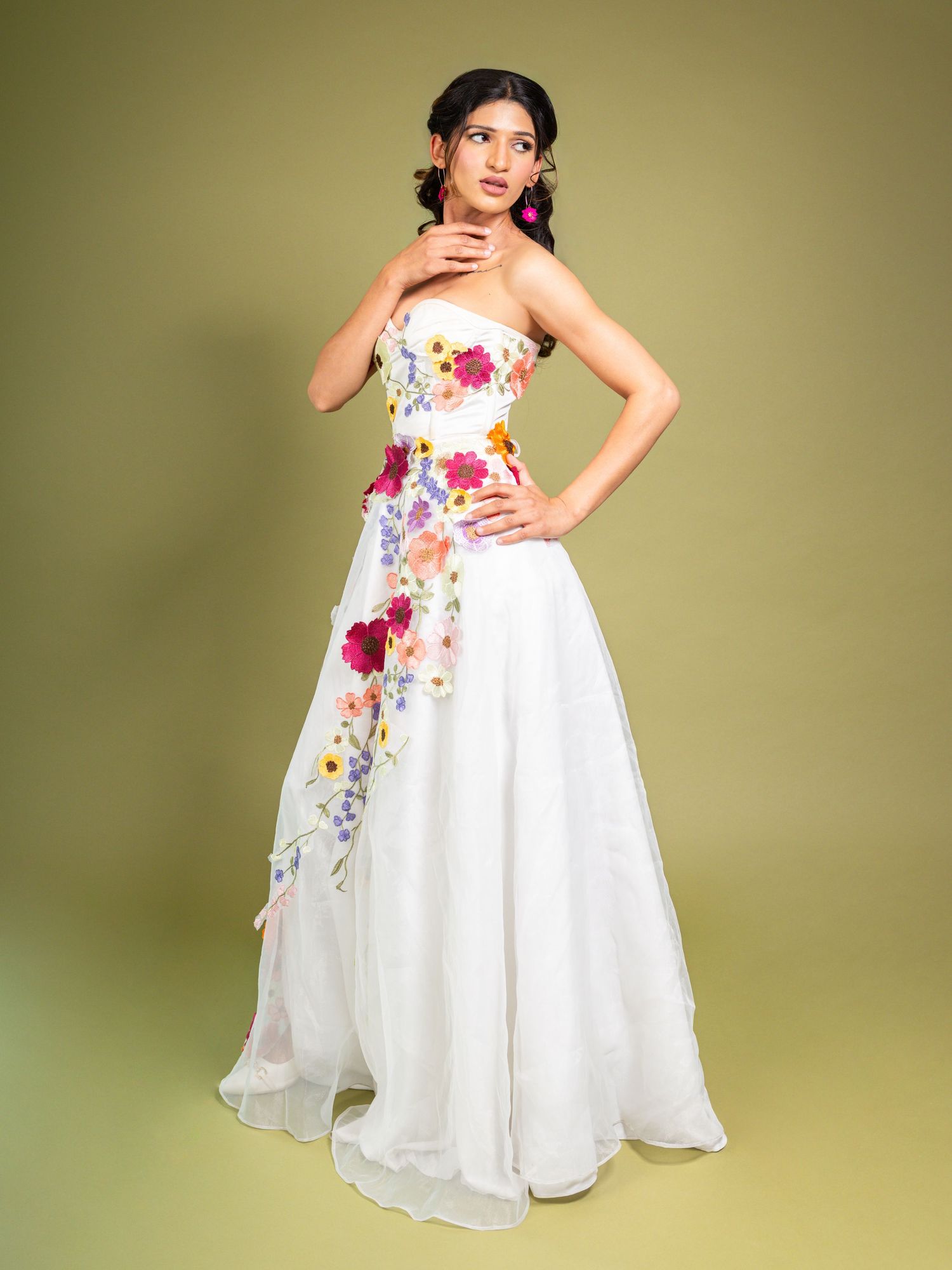 Woman in white strapless floral gown with colorful flower appliques, posing against green background.
