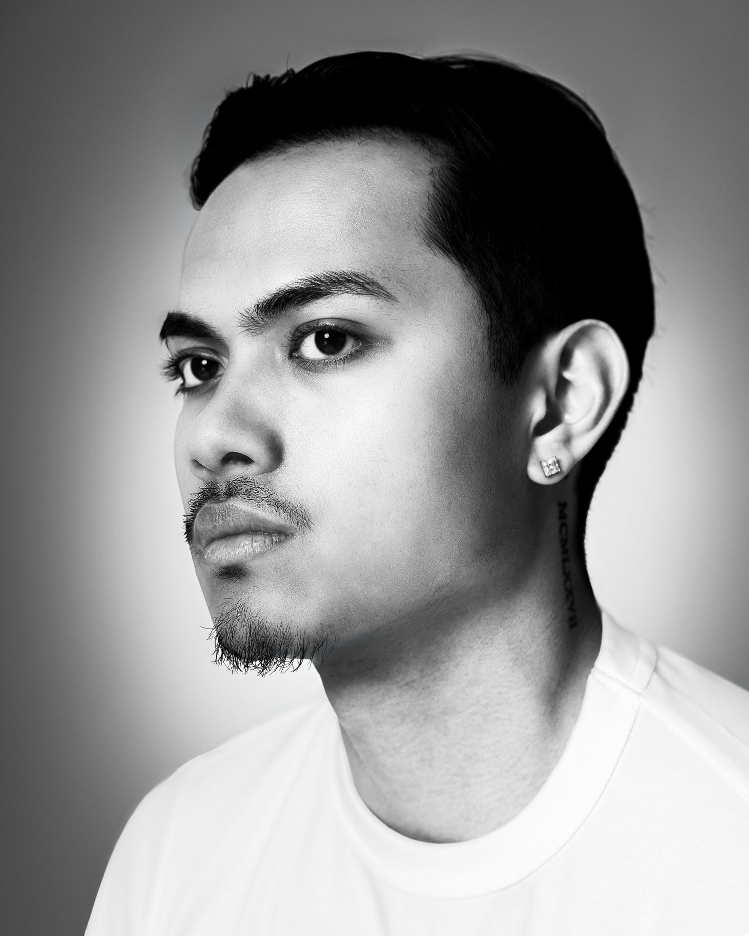 Black and white portrait of a young man with a small beard, earring, and short dark hair wearing a white t-shirt.