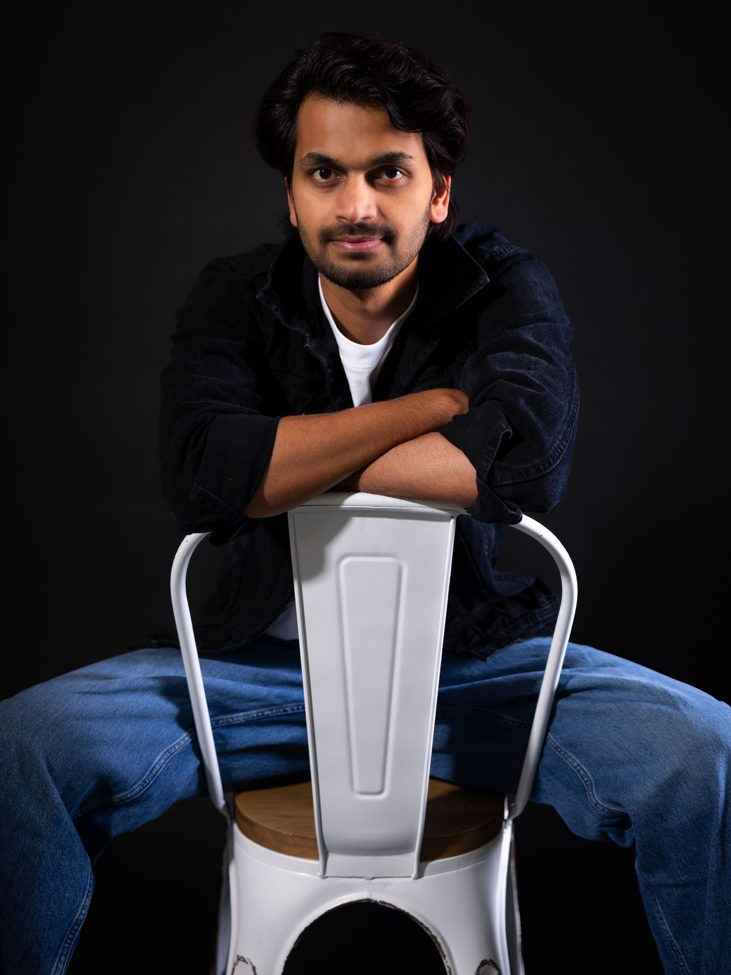 Young man in black jacket and jeans sits relaxed on white metal chair against dark studio background.