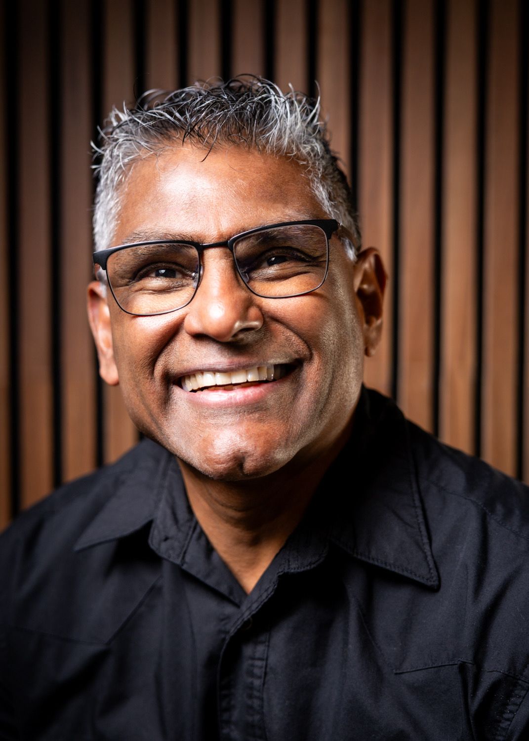 Smiling middle-aged man with gray hair and glasses wearing a black shirt against a wooden slat background.