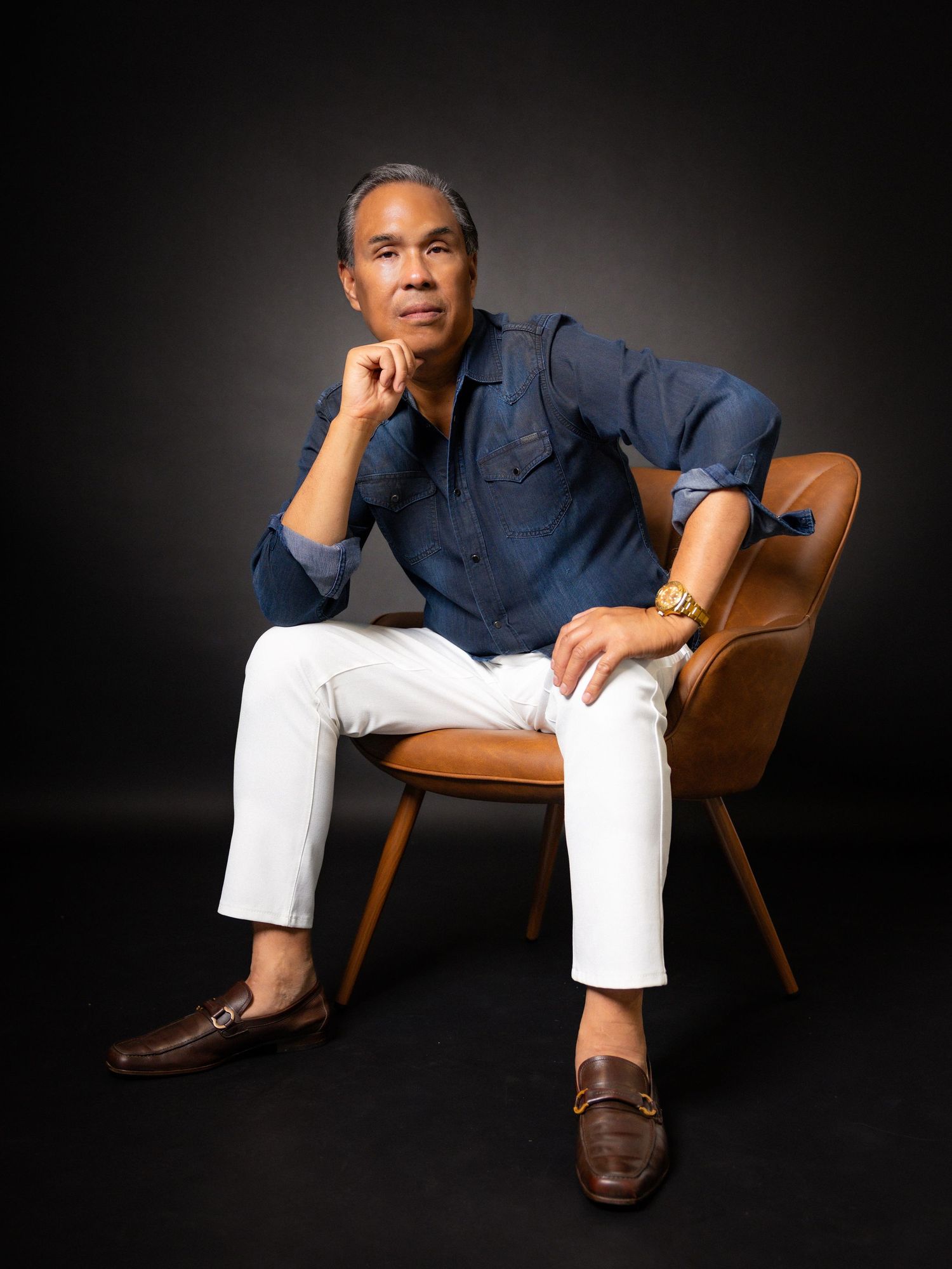 Middle-aged man in white pants and denim shirt sits thoughtfully in a leather chair against dark background.